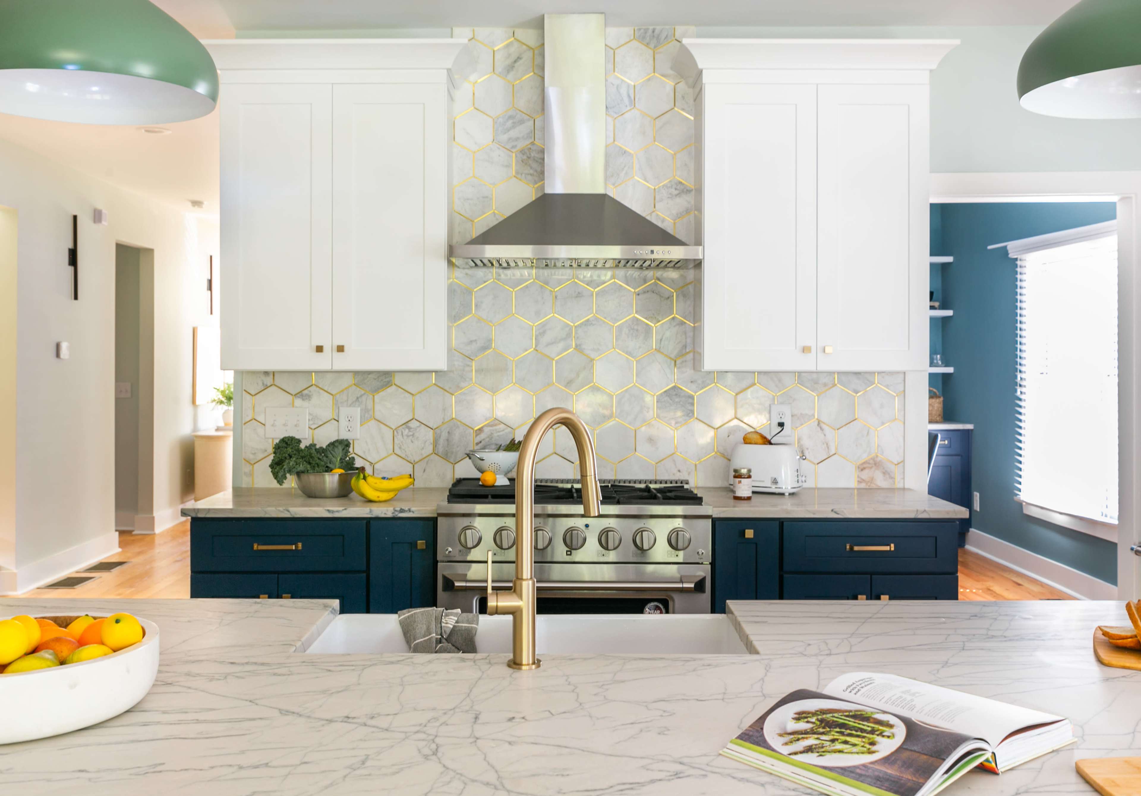 A modern kitchen featuring a marble countertop, a hexagonal tile backsplash, a stainless steel stove with a chimney hood, and two green pendant lights above an island with a bowl of oranges.