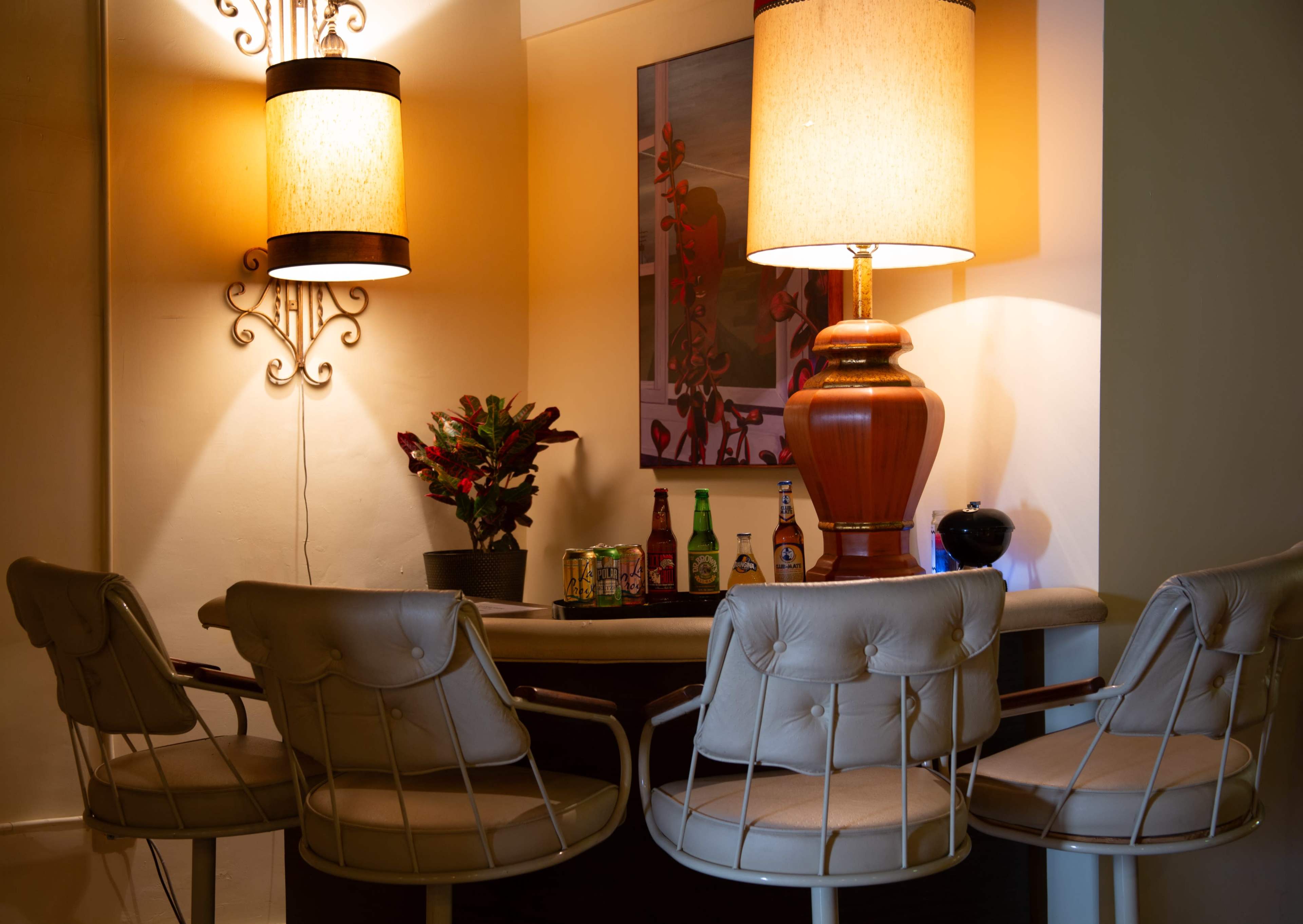A small round table is set in a softly lit corner, surrounded by four beige chairs, with drinks and a plant on the table and artwork on the wall.