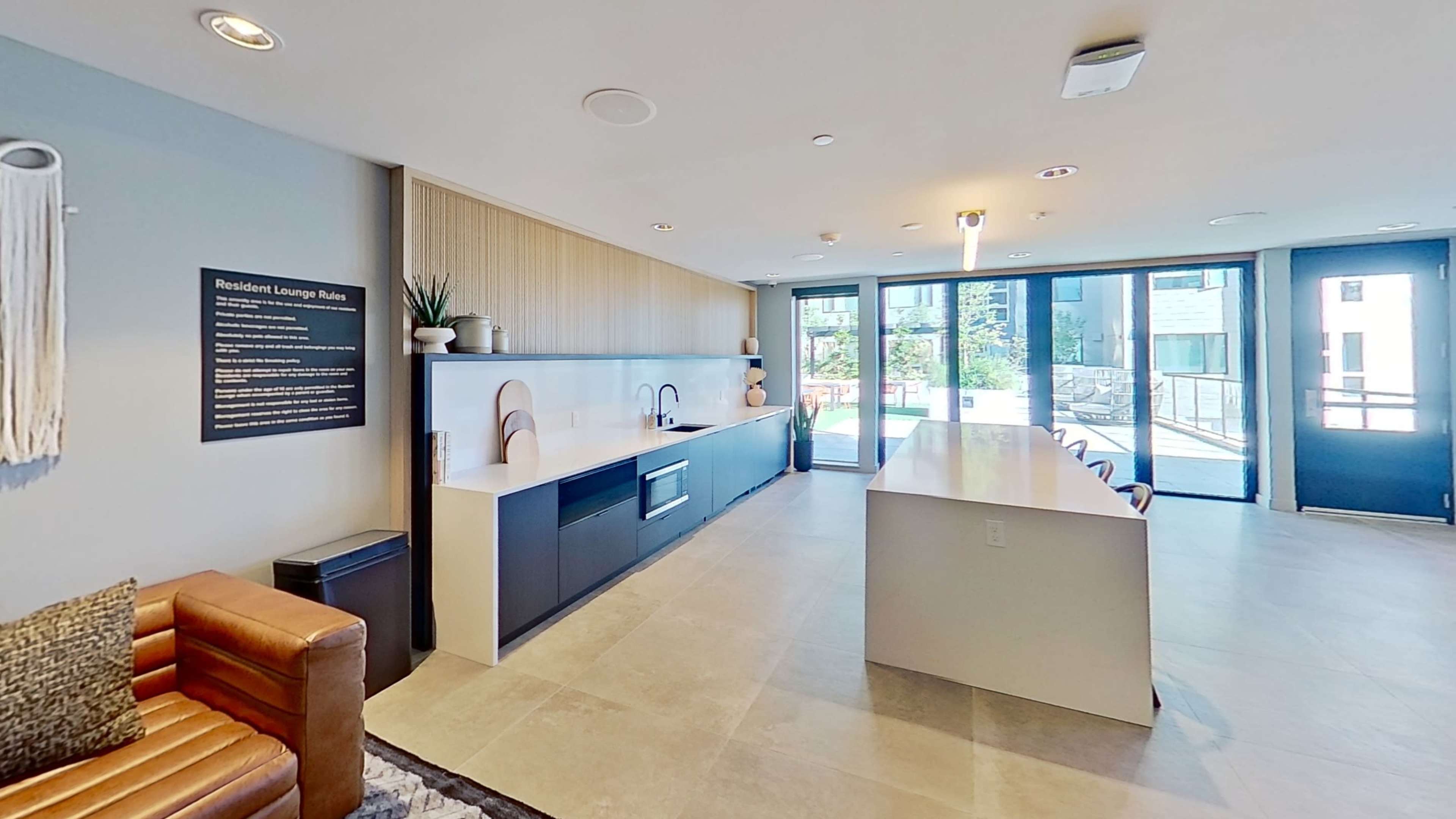 The image shows a modern lounge area with a kitchen counter, seating, and large glass doors leading to an outdoor space.