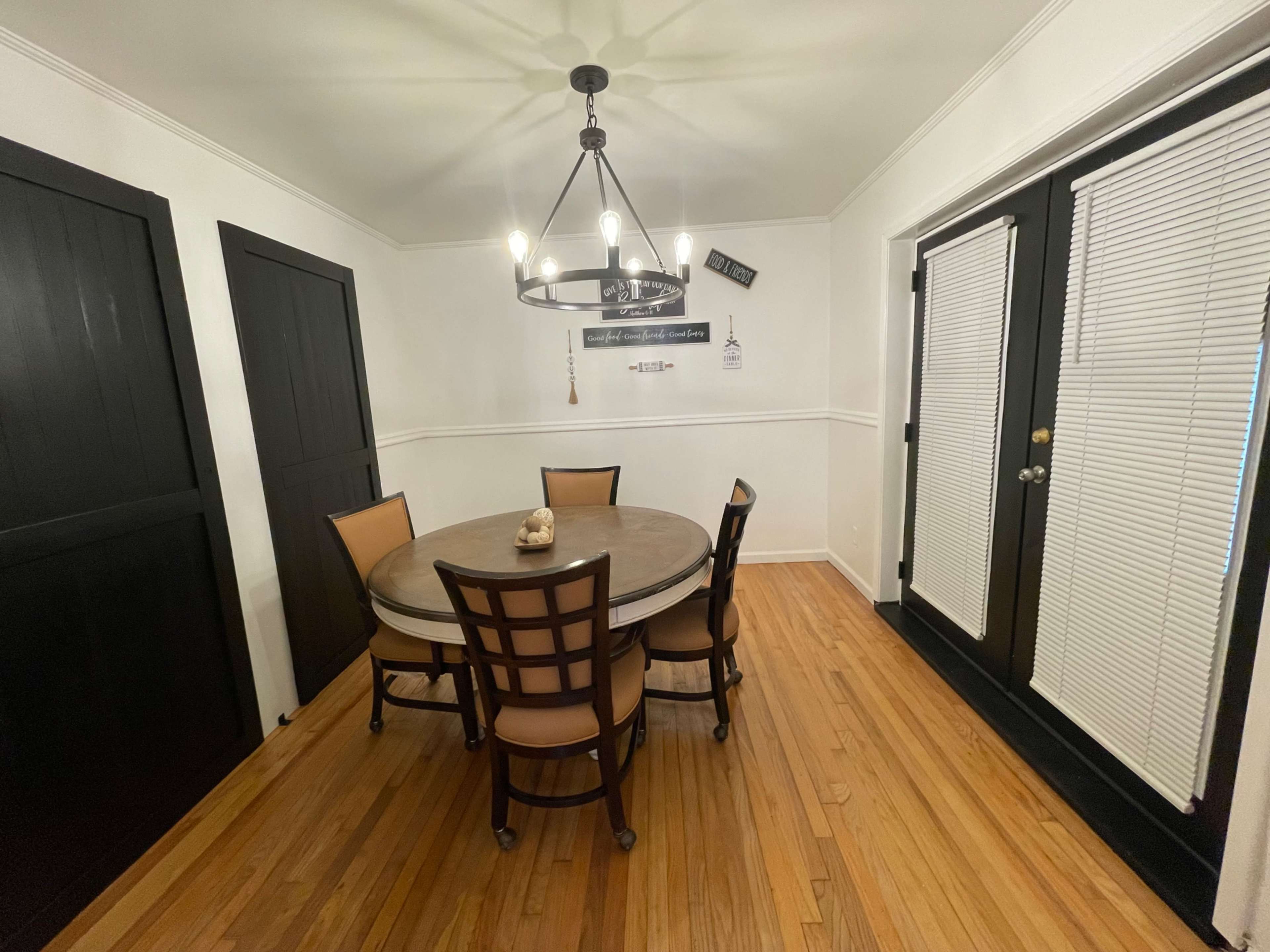 The image shows a dining room with a round wooden table and six chairs, accompanied by two dark doors and a large window with blinds.