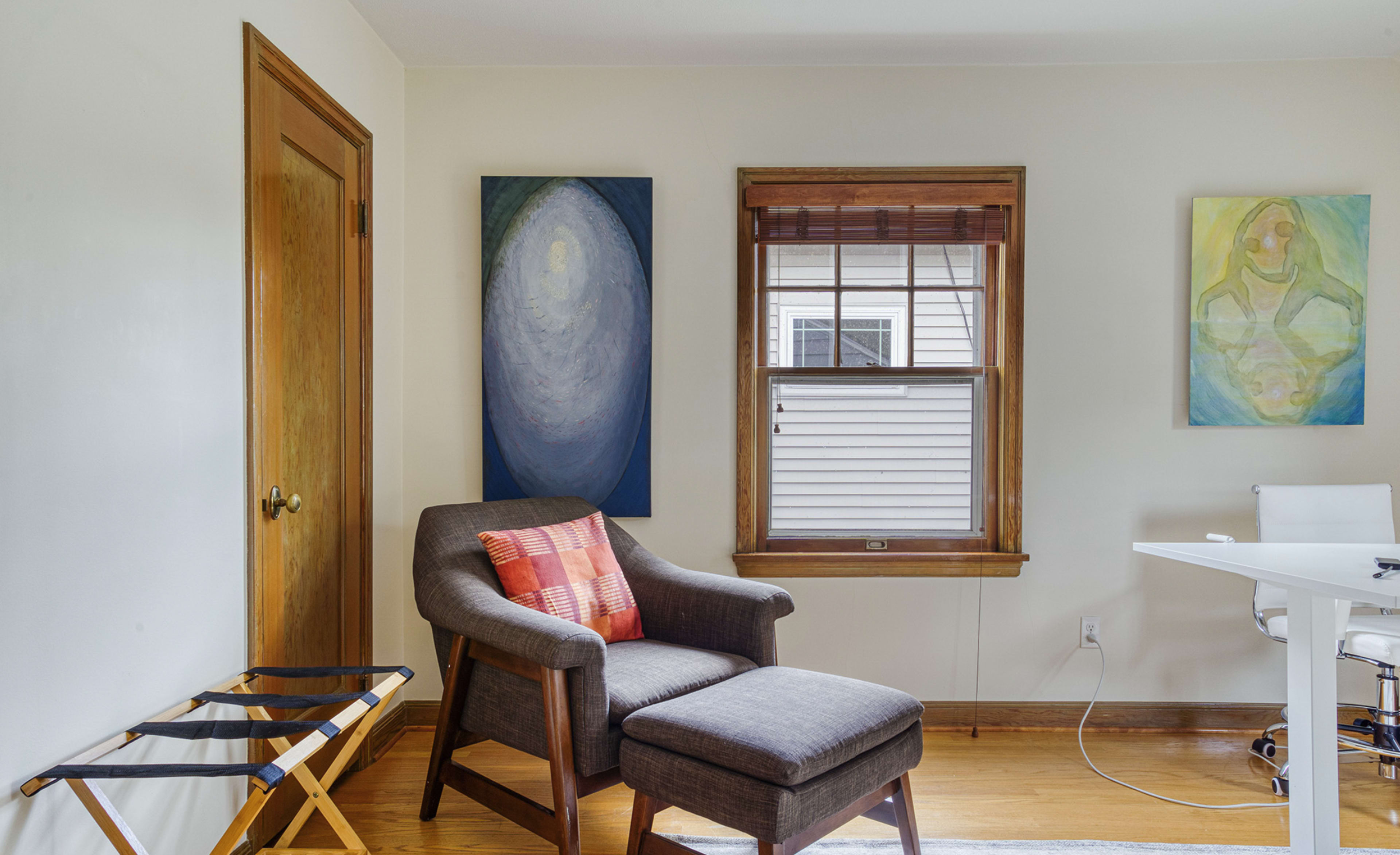 The image shows a cozy room featuring a gray armchair with a red plaid throw pillow, a small folding chair, two abstract paintings on the walls, and a desk with a white chair.