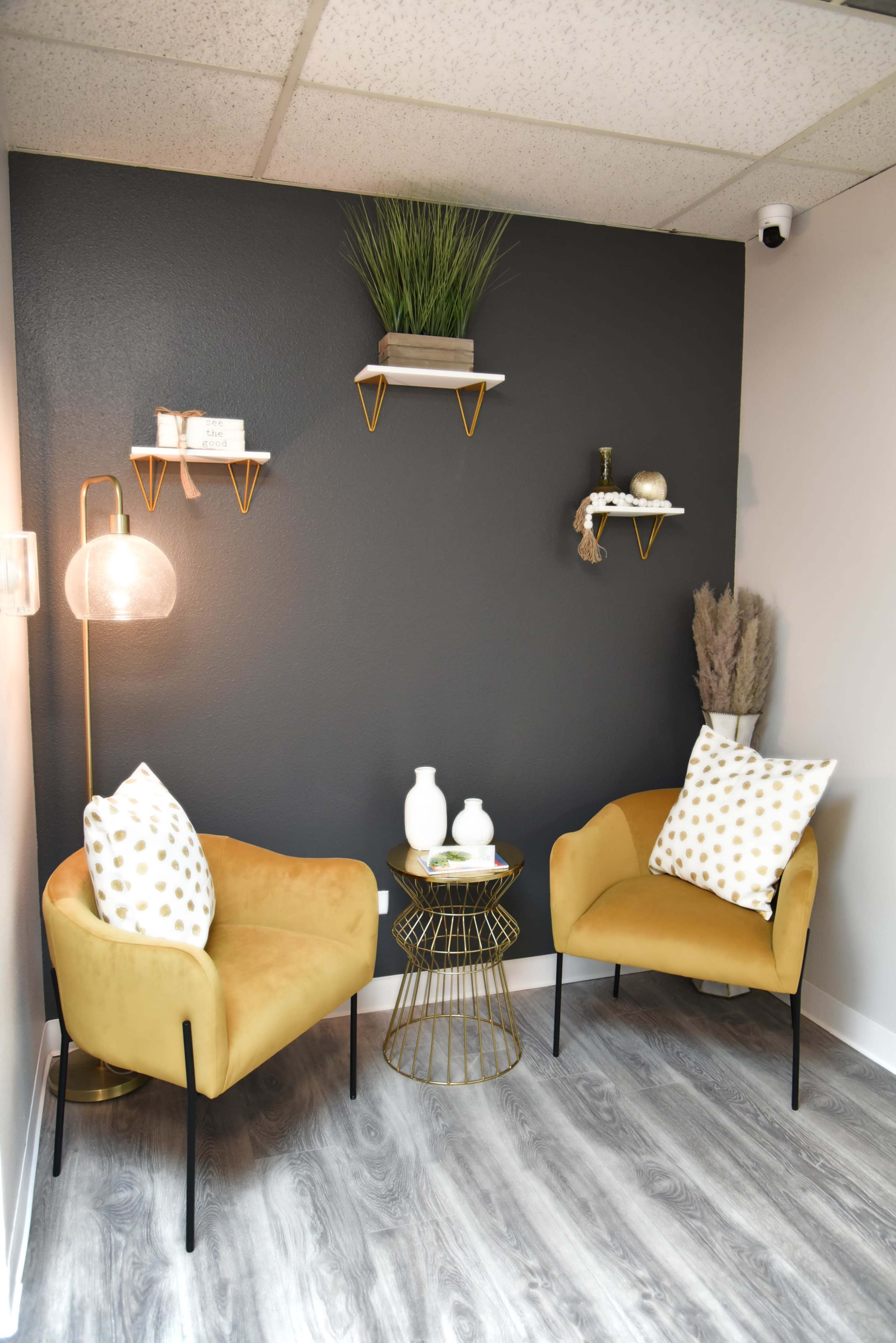 A cozy corner features two yellow chairs with decorative pillows, a small round table, and wall shelves displaying plants and decor against a dark wall.