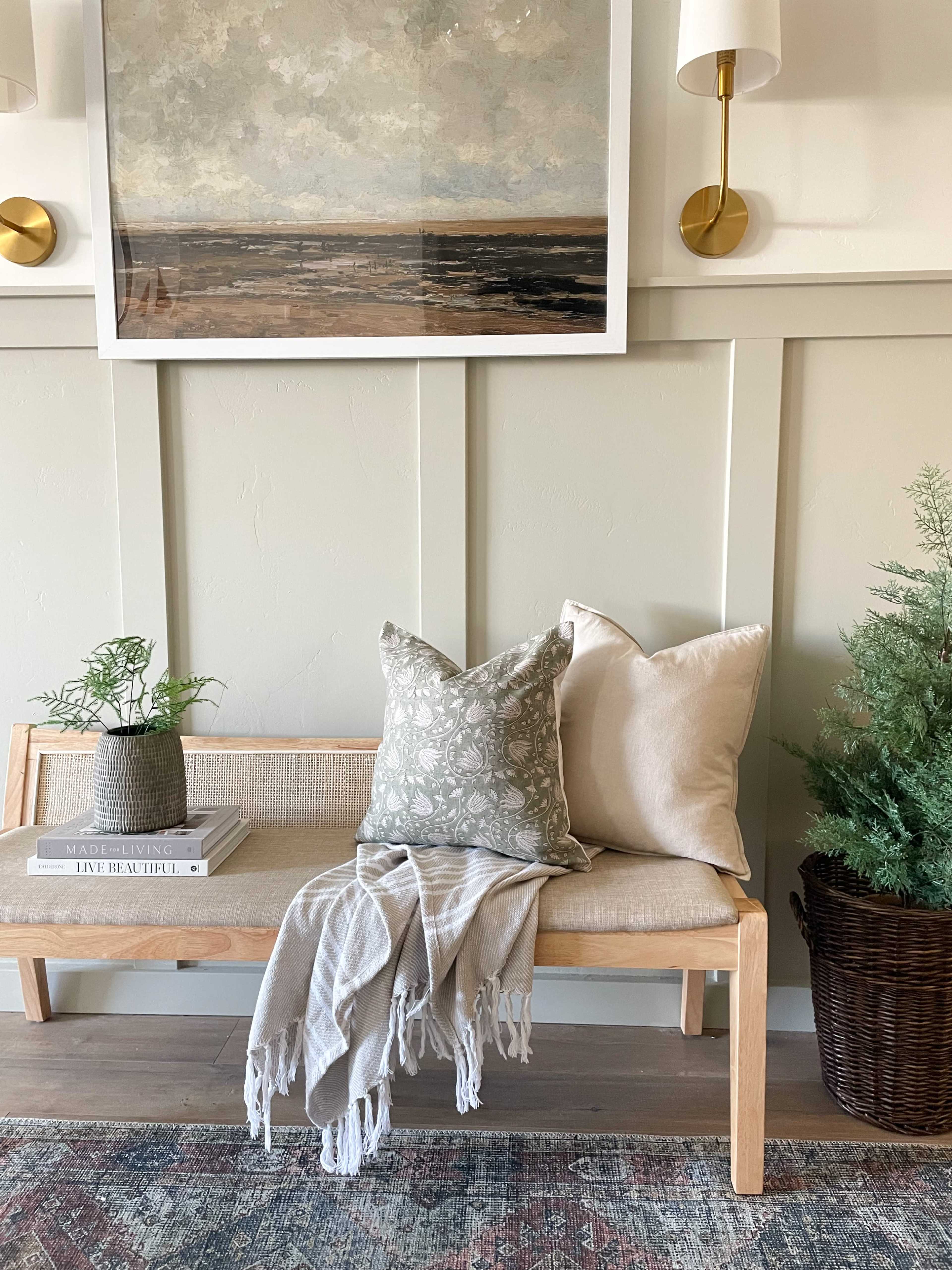 A wooden bench is adorned with two decorative pillows, a textured throw blanket, and a potted plant, set against a light-colored wall featuring framed artwork and wall sconces.