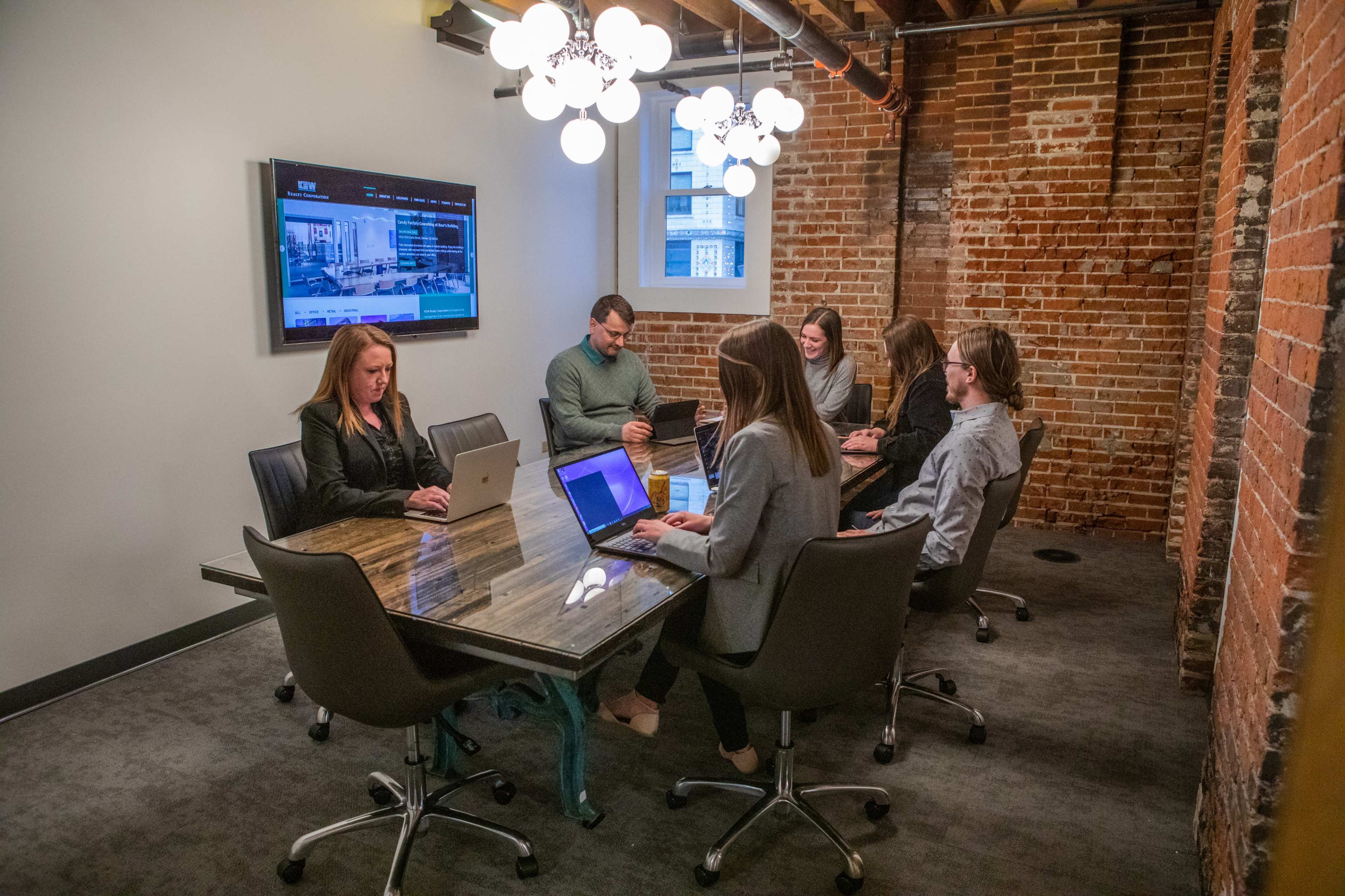 A group of six people, seated around a conference table, works on laptops in a meeting room with exposed brick walls and modern lighting.