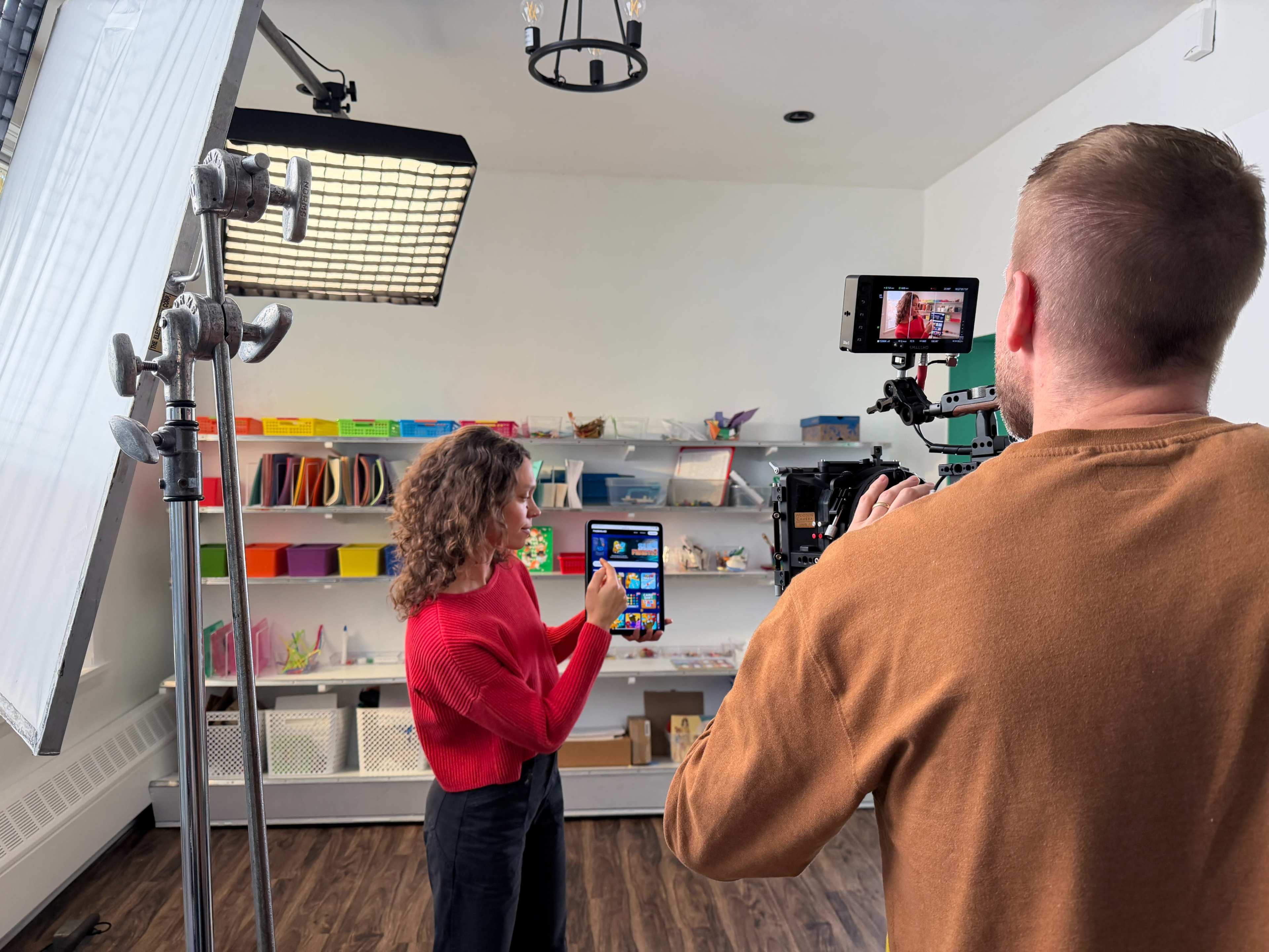 A woman in a red sweater gestures while being filmed by a camera operator in a studio filled with colorful school supplies in the background.