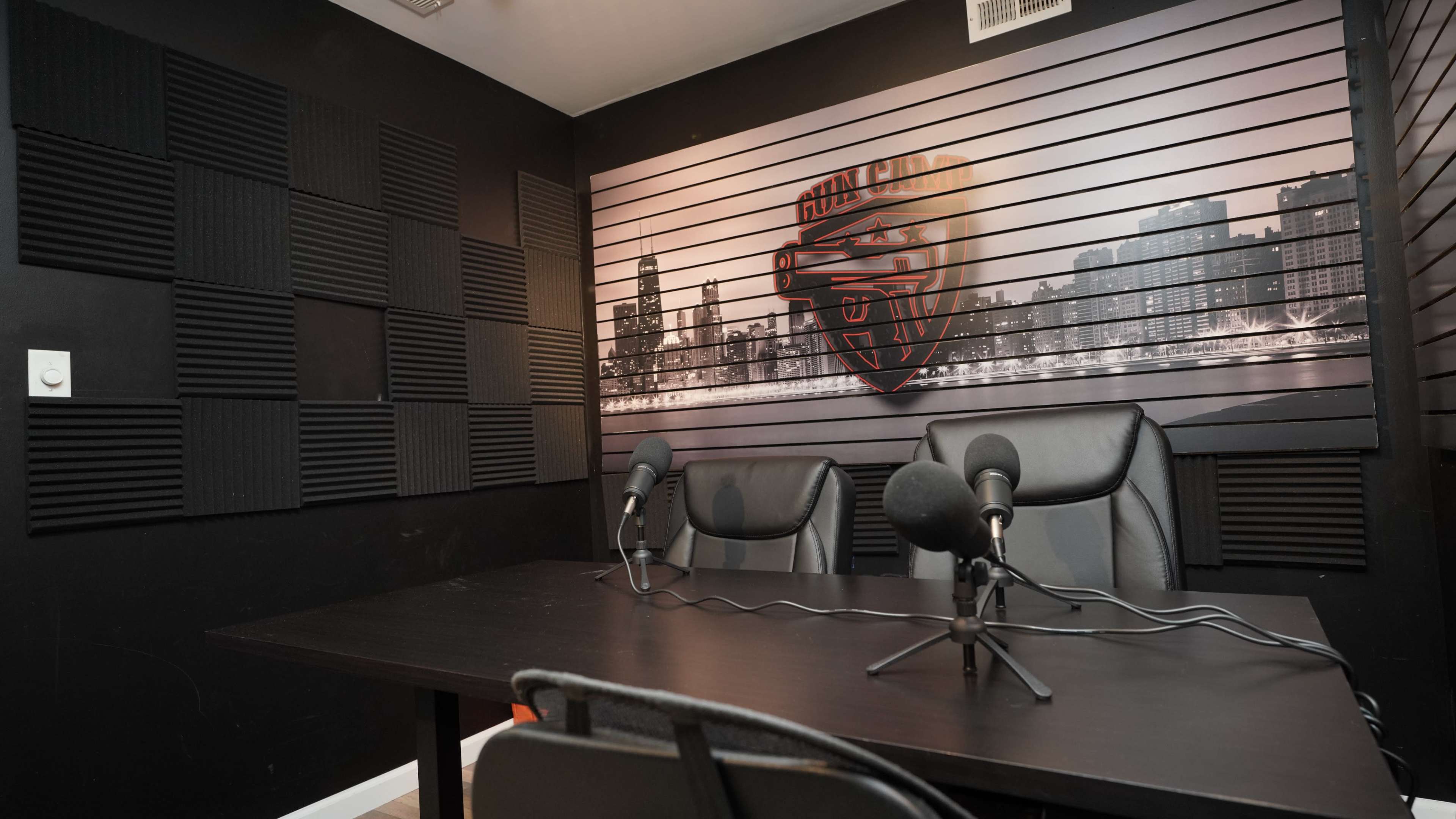 A podcast studio with a black table, two microphones, and a wall featuring a graphic of a city skyline and a logo.