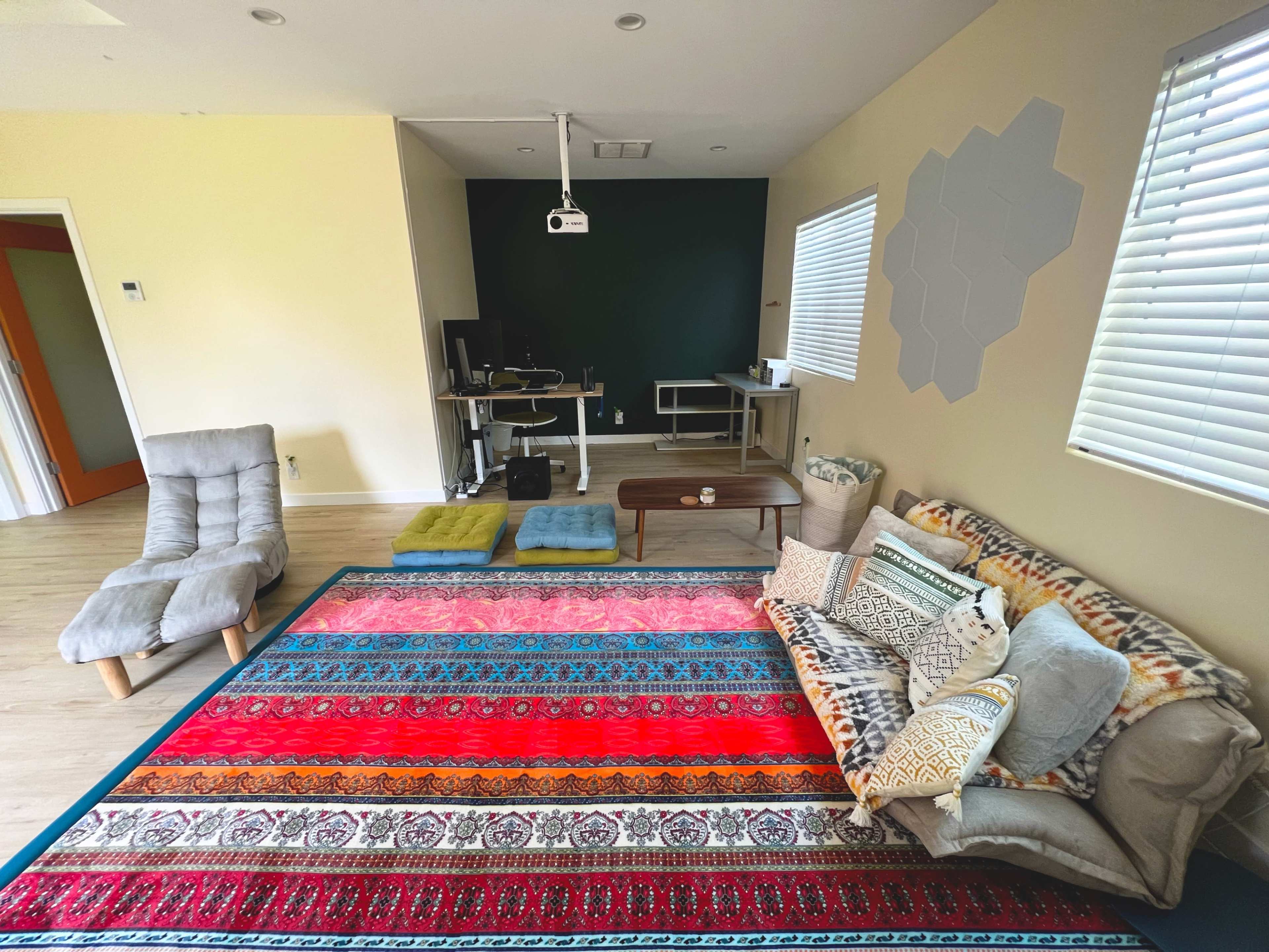 The room features a colorful area rug, a sofa with various cushions, a low coffee table, and a workspace with a computer setup against a green accent wall.
