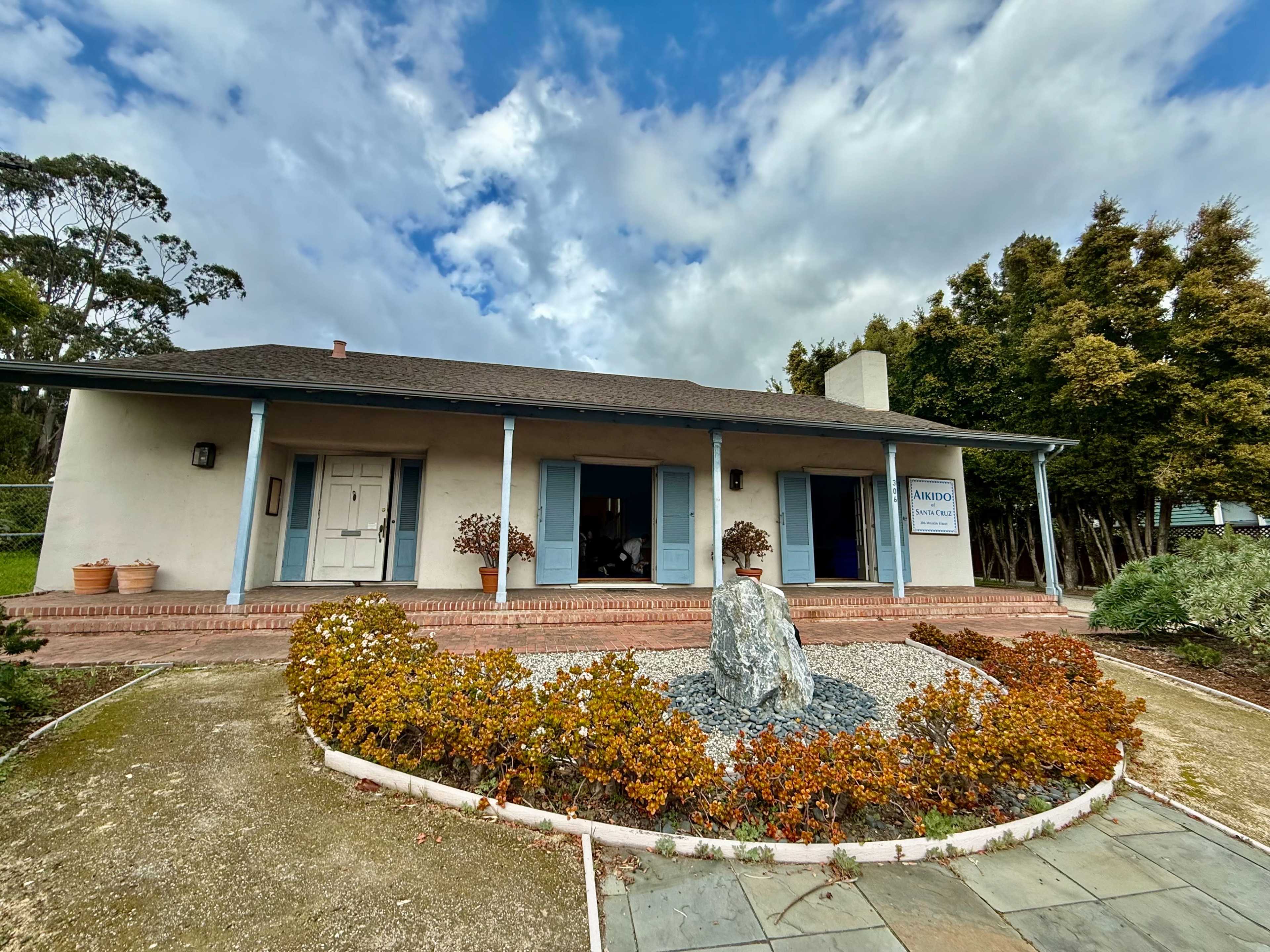 A one-story building with a planted garden and a large rock in front is set against a cloudy sky.