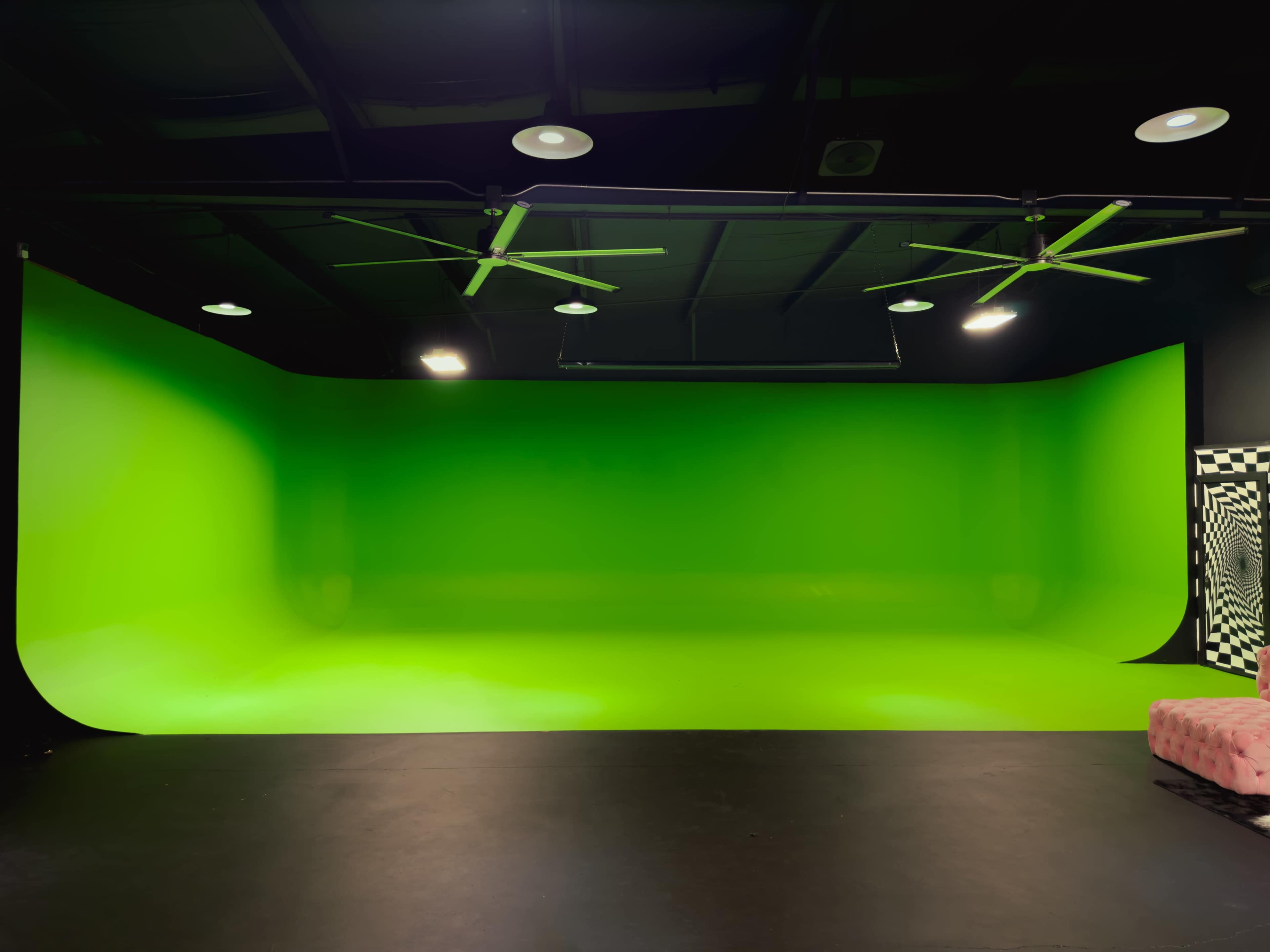 A studio with a bright green backdrop and a black floor, illuminated by ceiling lights and fans.