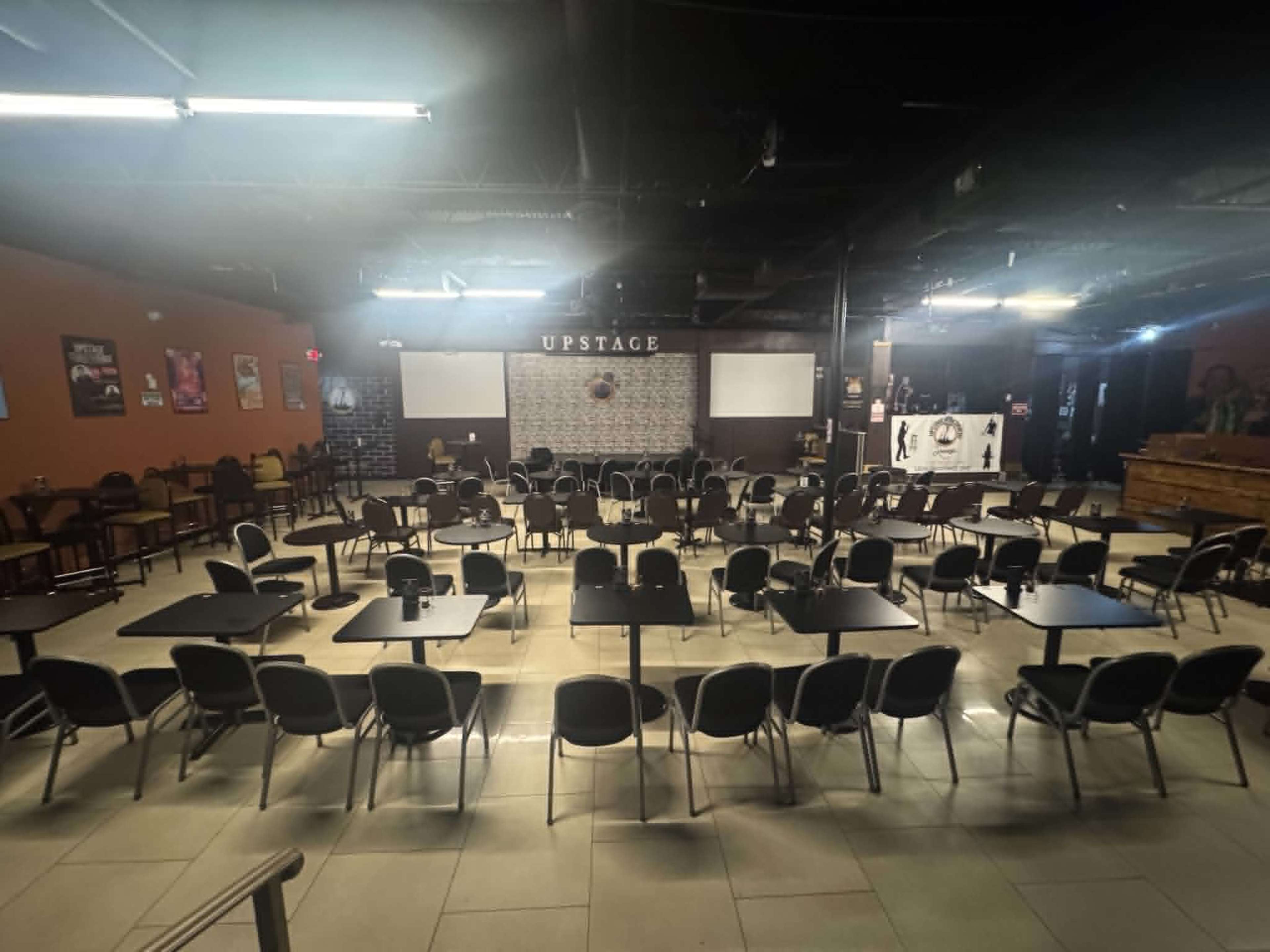 The image shows a spacious venue with rows of tables and chairs arranged neatly for an event, featuring a stage in the background labeled "UPSTAGE."