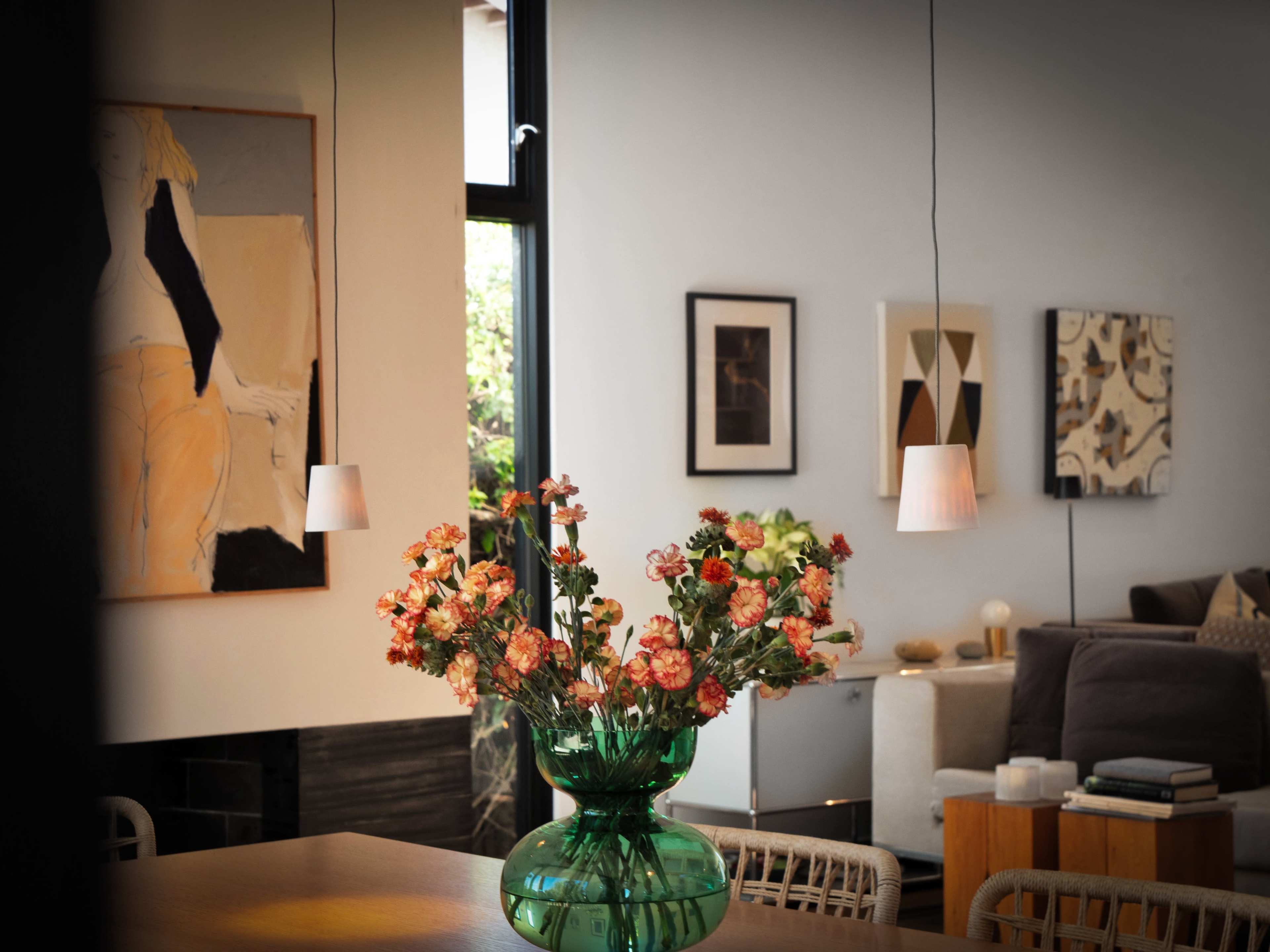 A green glass vase filled with flowers is placed on a table in a modern living space featuring art on the walls and pendant lights overhead.