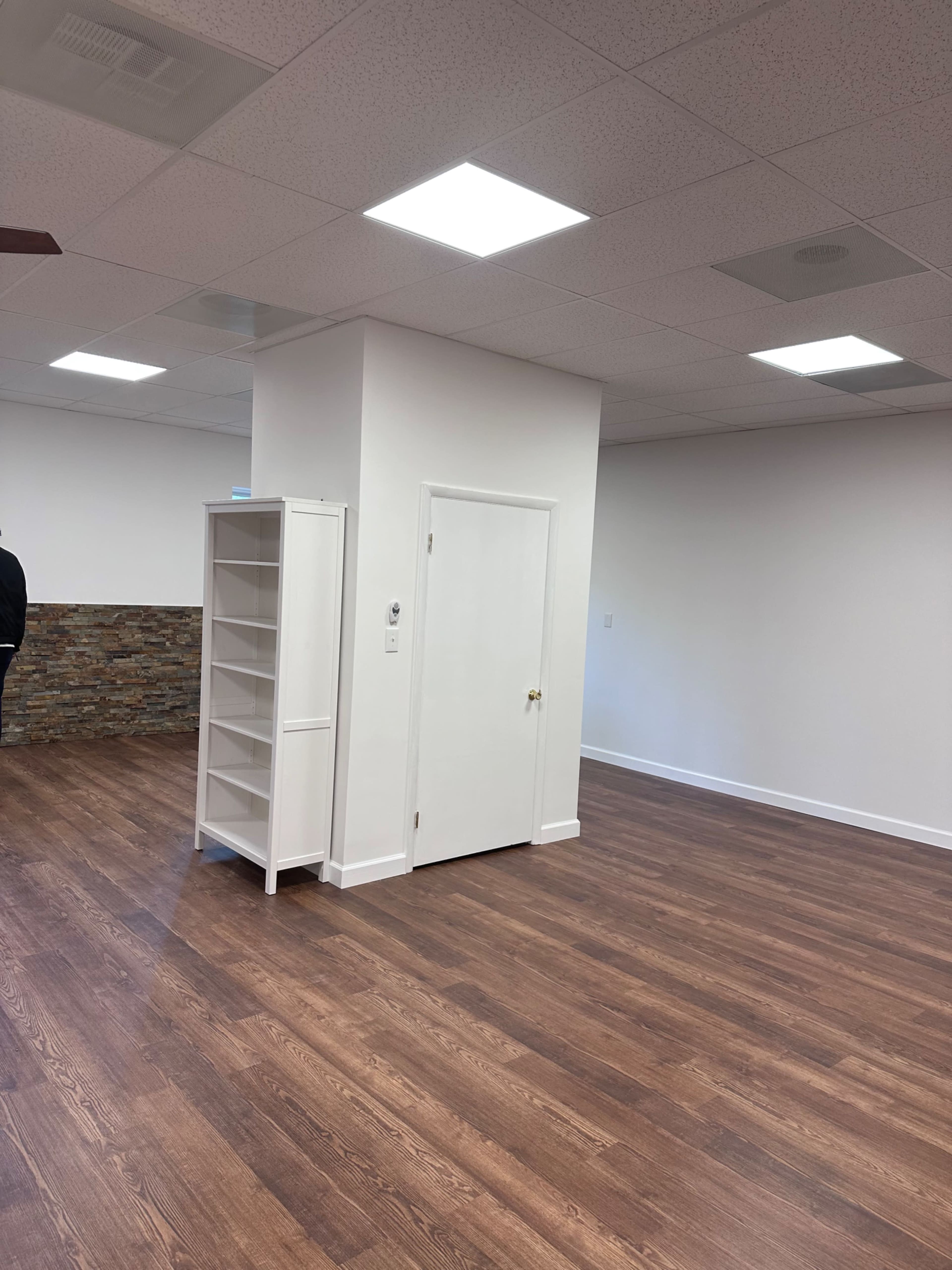 The image shows a spacious, well-lit room with wooden flooring, featuring a corner shelf unit and a doorway.
