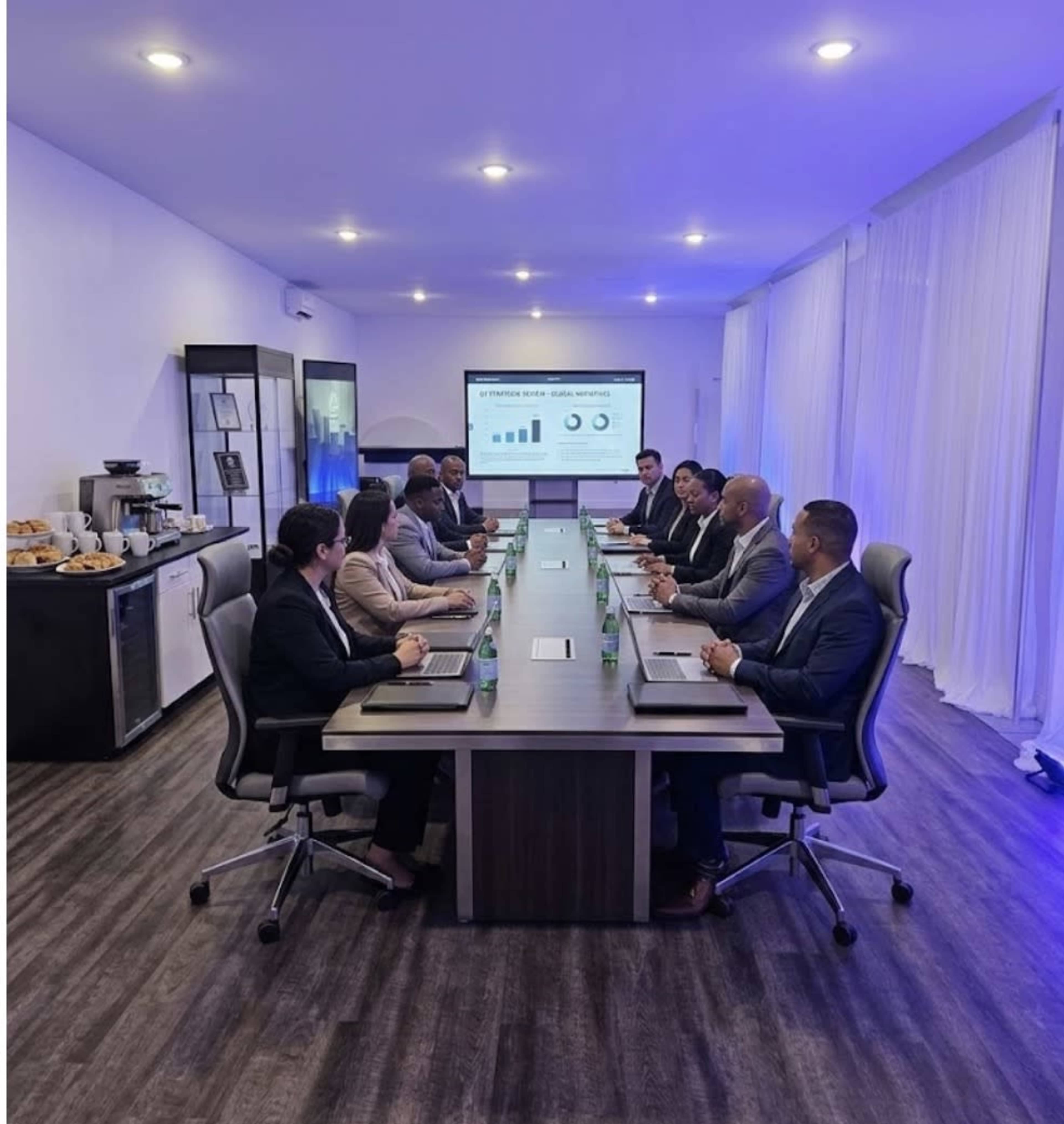 A group of eight professionals is seated around a long conference table in a modern meeting room, while a presentation displaying graphs is projected on a screen at the front.