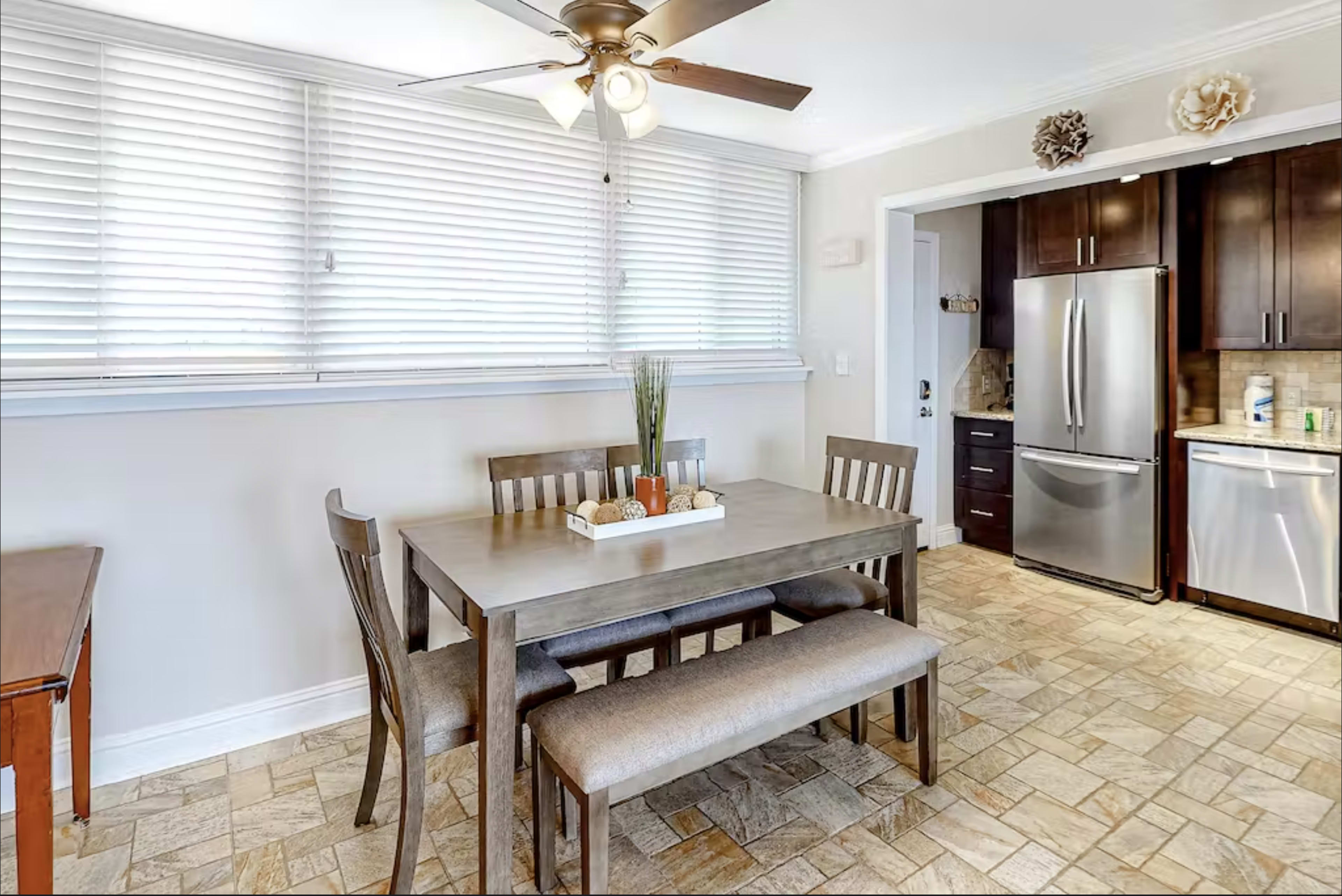 A dining area features a wooden table with benches, surrounded by light-colored walls and a kitchen with stainless steel appliances in the background.