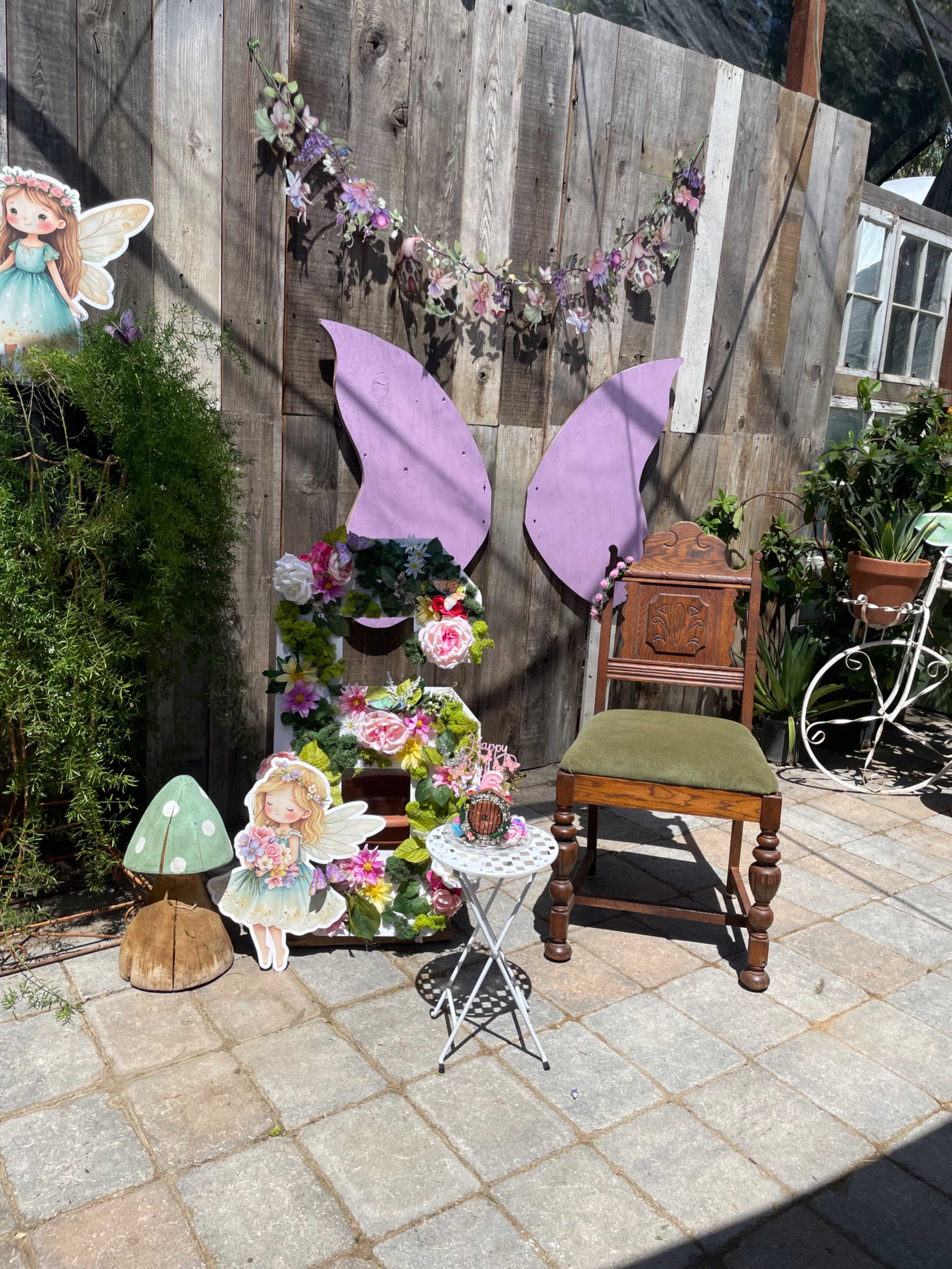 The image shows a whimsical corner setup with a wooden wall, decorated with fairy-themed elements, including large purple wings, flowers, and a small mushroom.