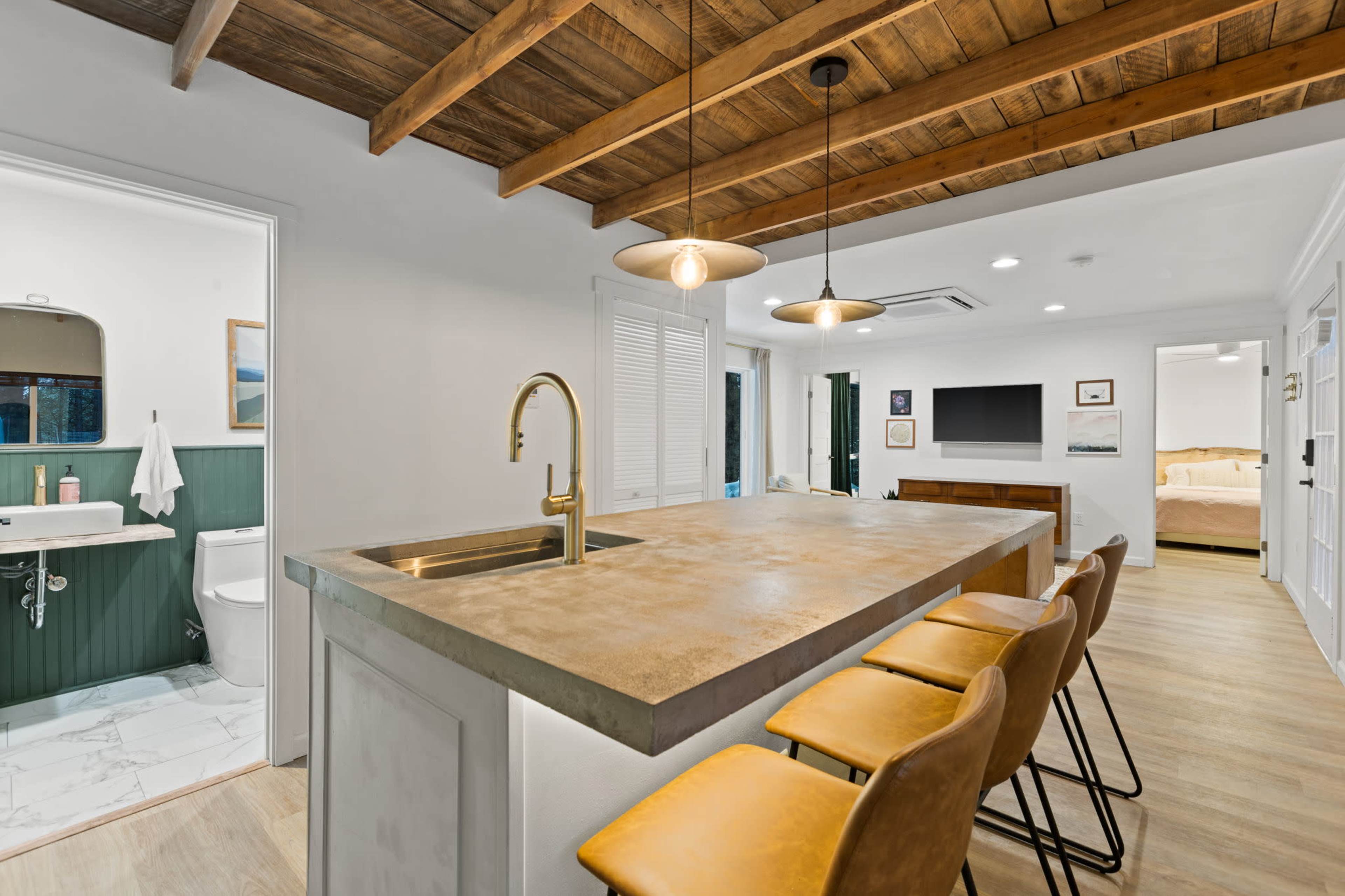 The image shows a modern kitchen with a concrete island, wooden beams overhead, and four yellow bar stools, adjacent to a bathroom and living area.