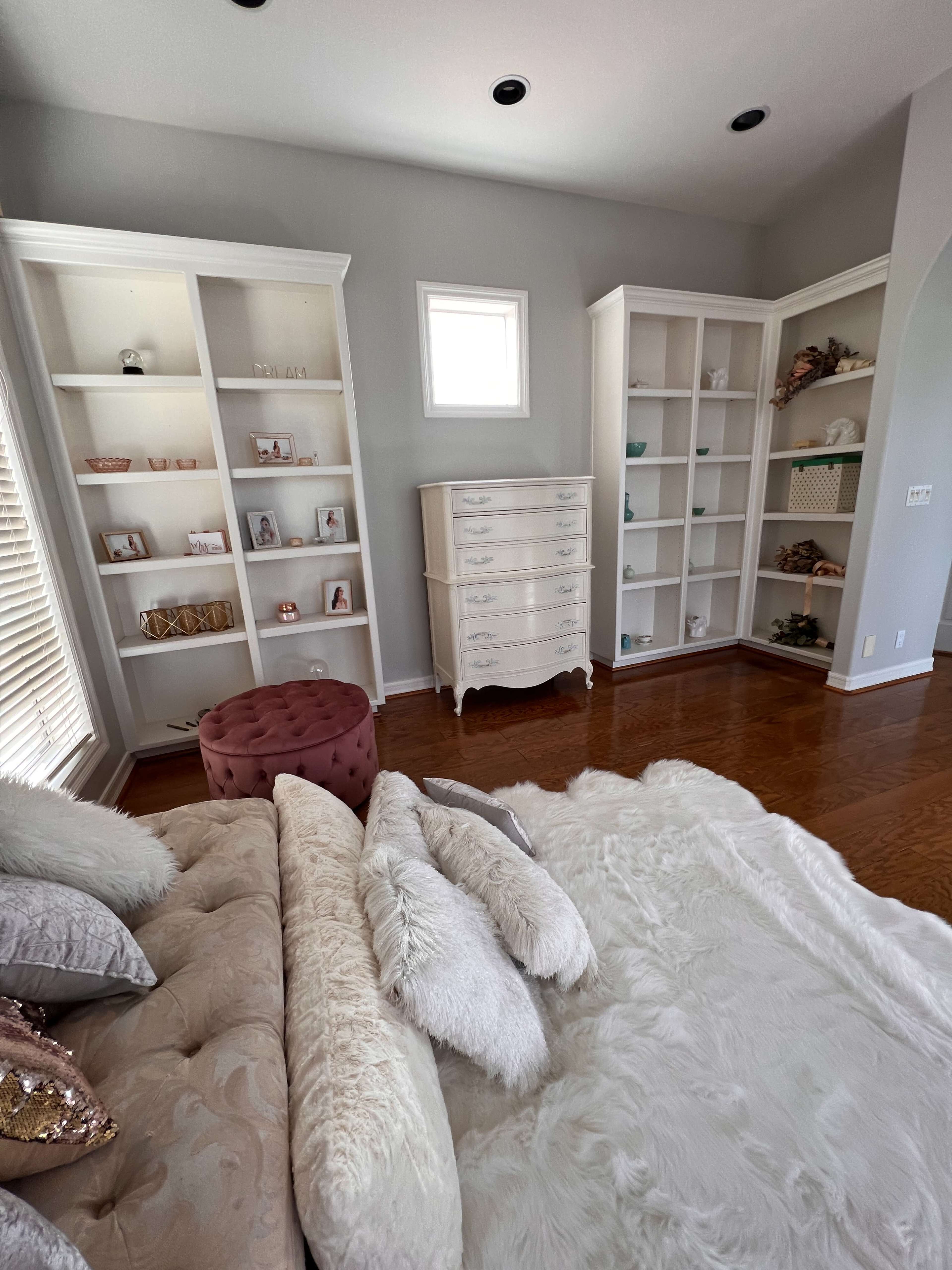 The image shows a well-lit living room with white built-in shelves, a light-colored dresser, a plush sofa, and a large area rug.