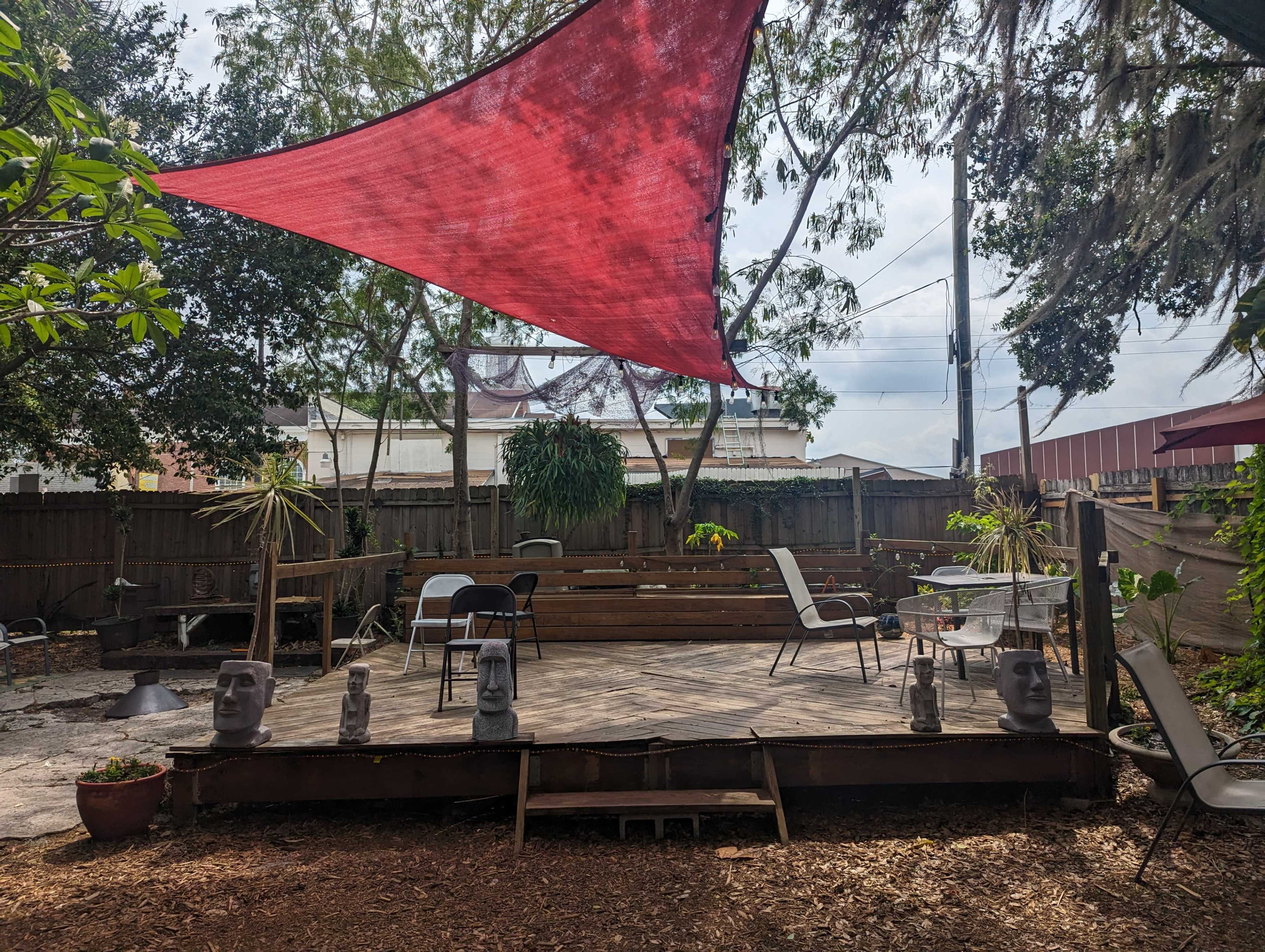 A backyard patio features a wooden deck with chairs, decorative statues, and a large red shade sail overhead.