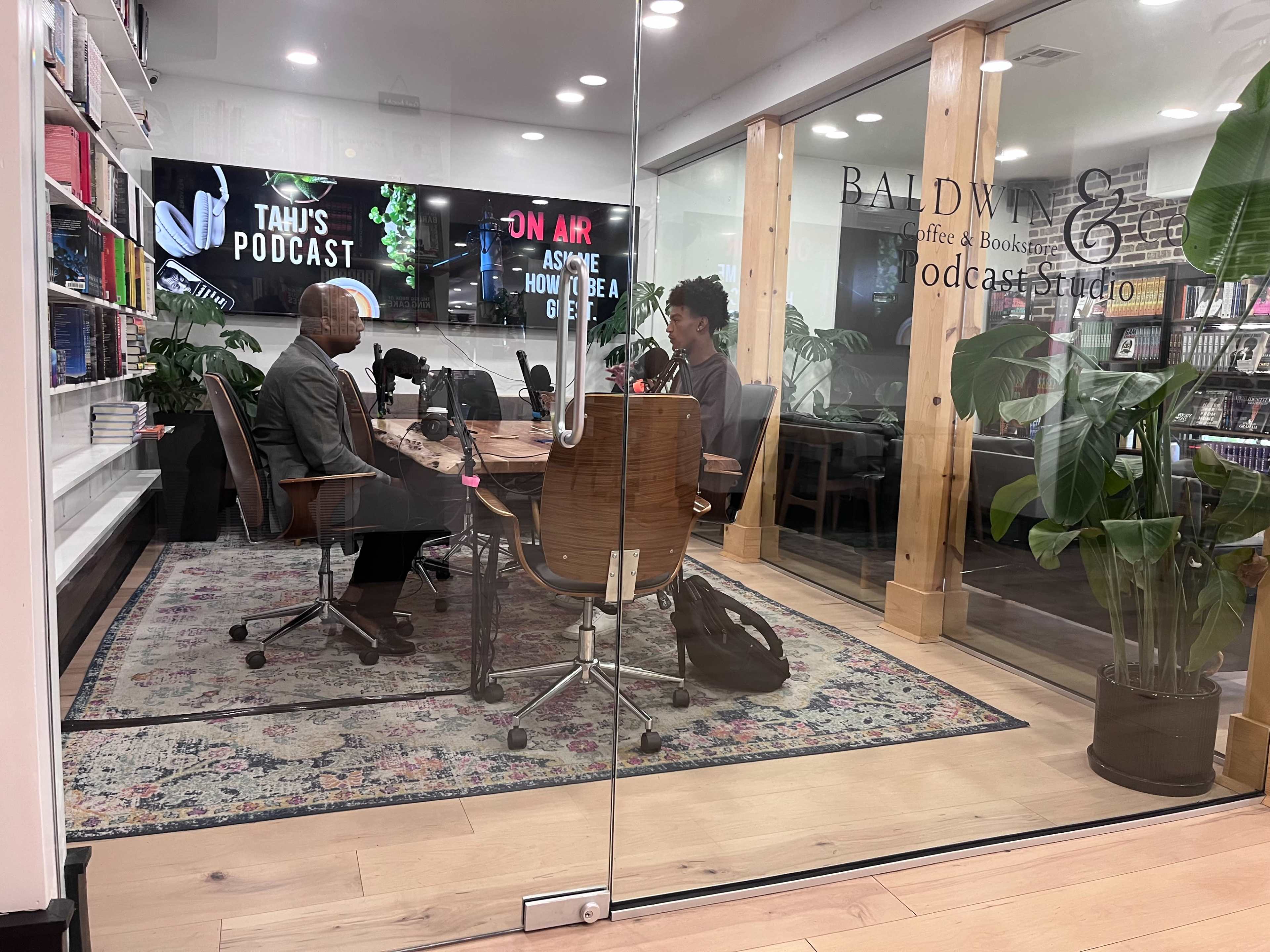 A man and a woman sit across from each other at a table in a podcast studio equipped with microphones and bookshelves.