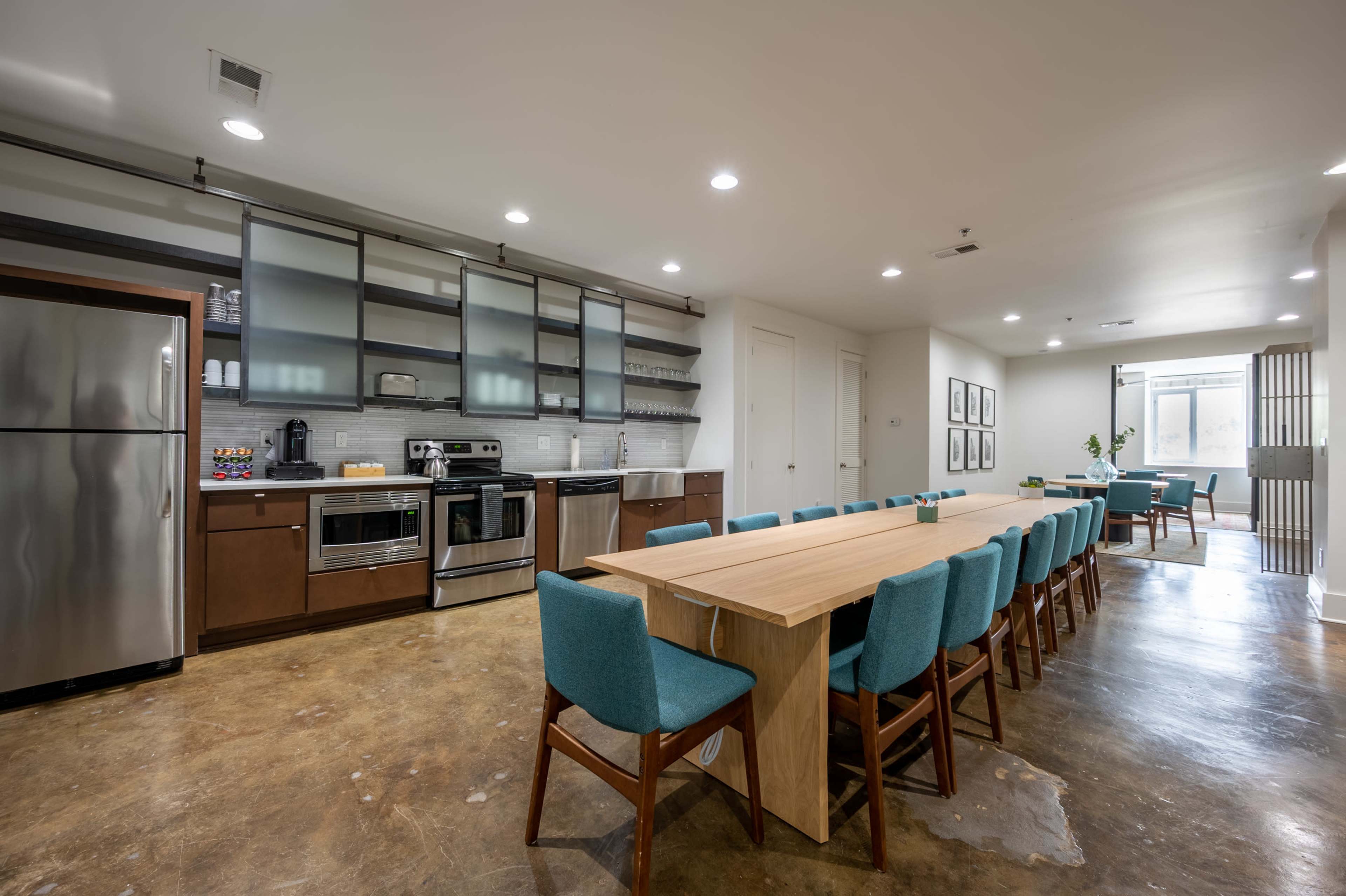 A modern kitchen and dining area featuring stainless steel appliances, a long wooden table with teal chairs, and a minimalist design.