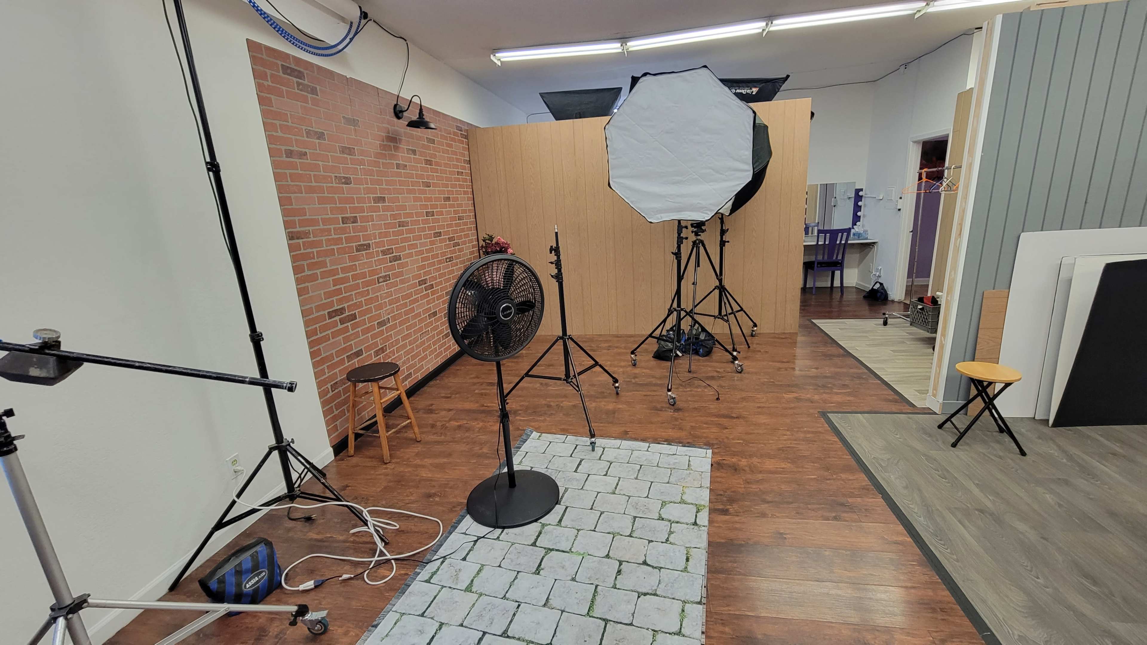 A photography studio with a brick wall backdrop, lighting equipment, a fan, and various types of furniture scattered around.