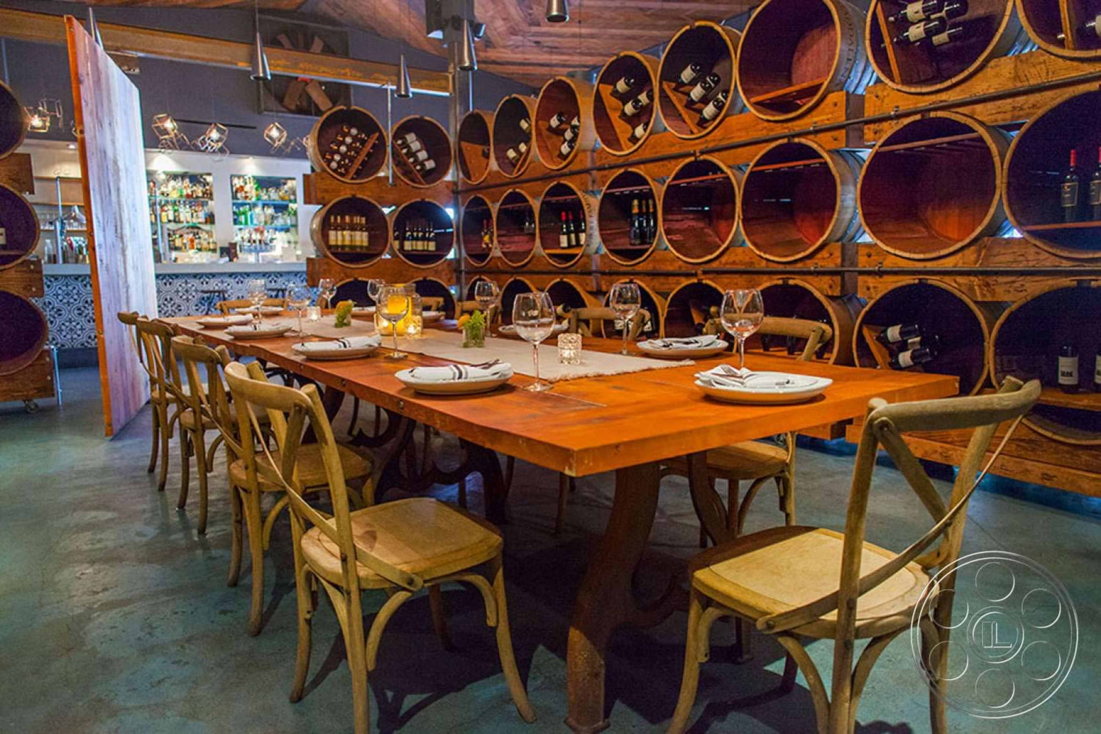 A wooden dining table is set with plates and glasses, surrounded by chairs, against a backdrop of stacked wine barrels in a restaurant.