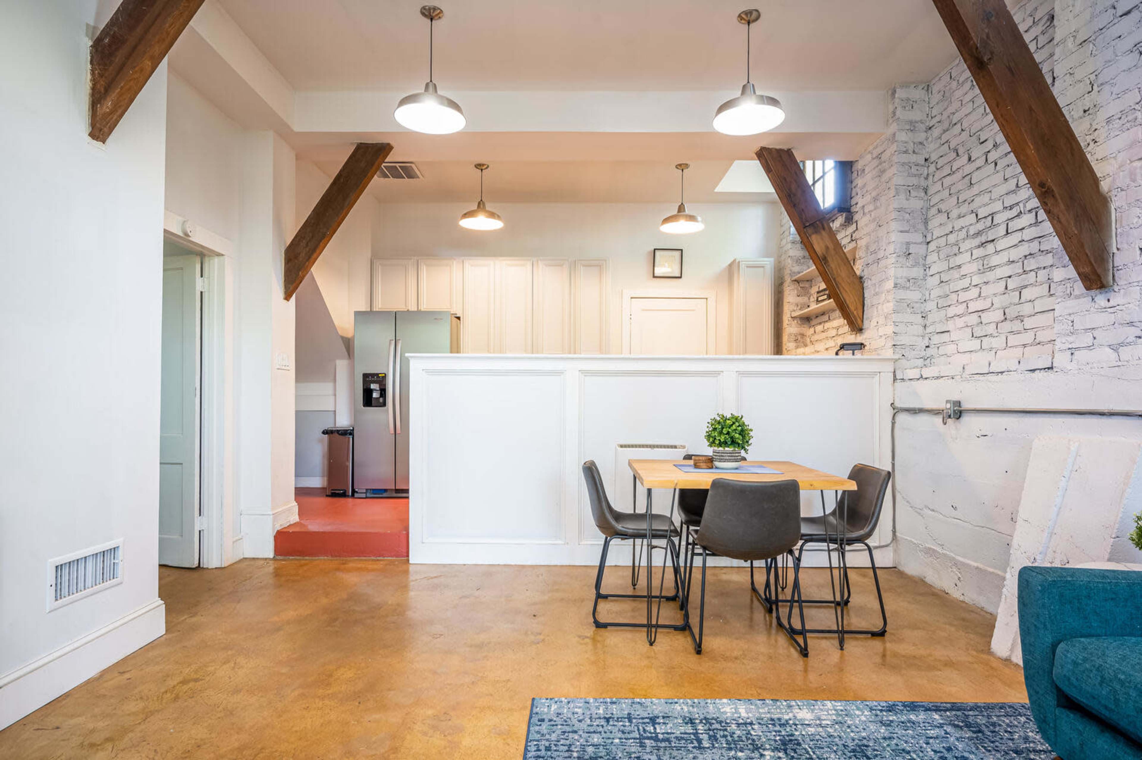 A modern living space featuring a dining area with a table and chairs, exposed wooden beams, and a kitchenette beyond a white partition.
