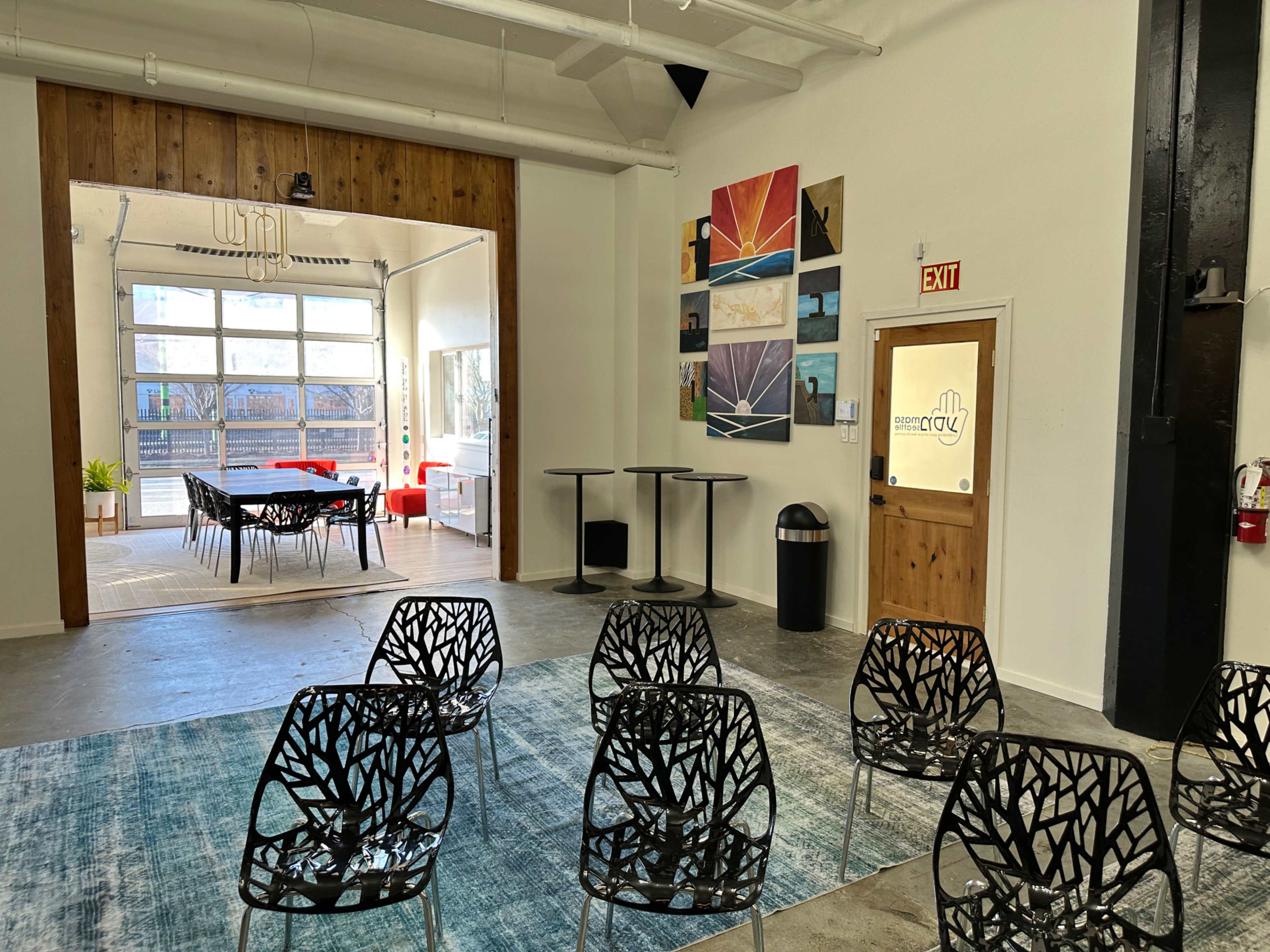 The image shows a spacious room with modern, black mesh chairs arranged in rows and a door leading to an area with tables and chairs set up for dining or meetings.