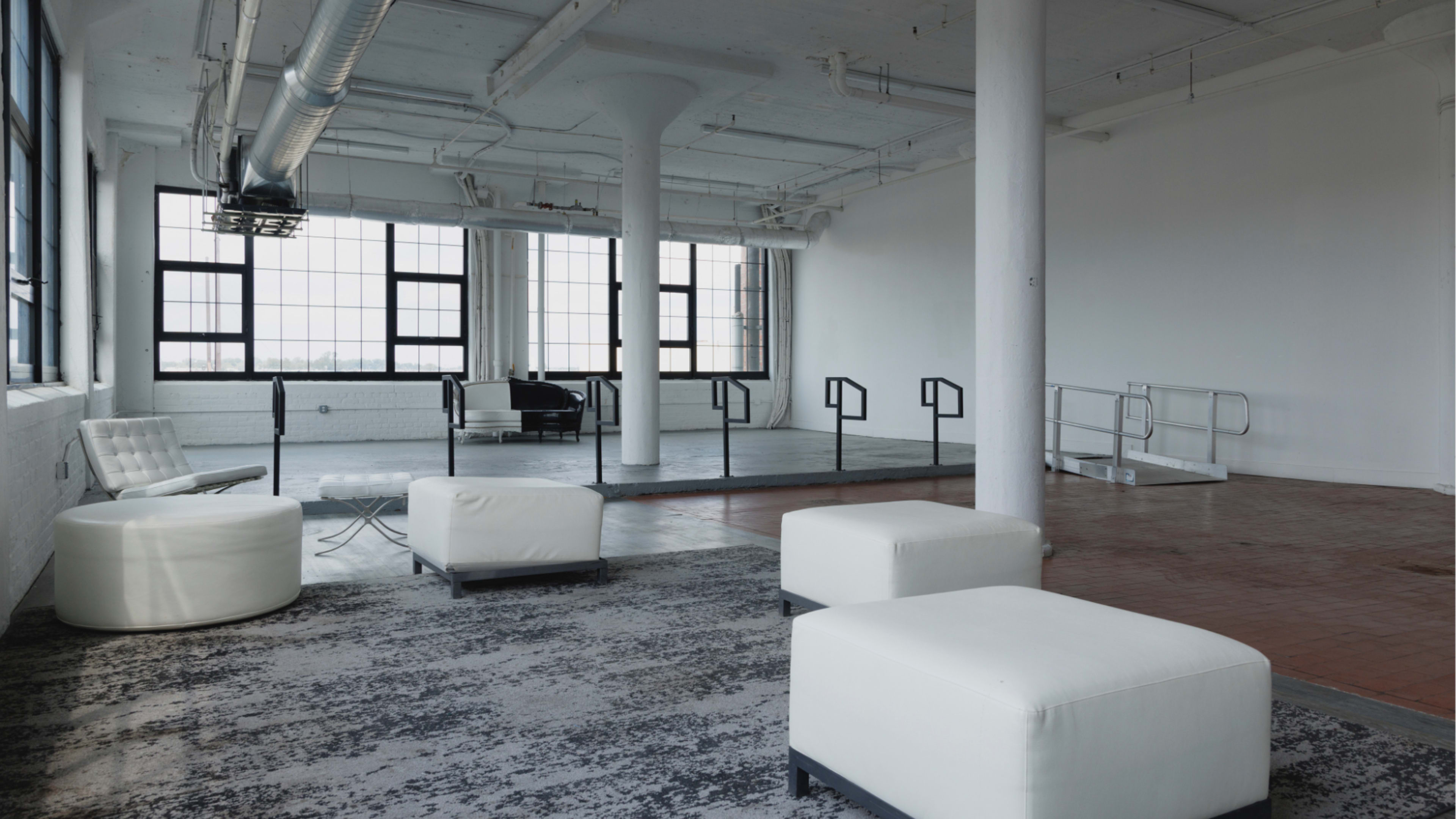 The image shows a spacious, empty industrial-style room with large windows, concrete pillars, and minimalist furniture set against a contrasting floor.