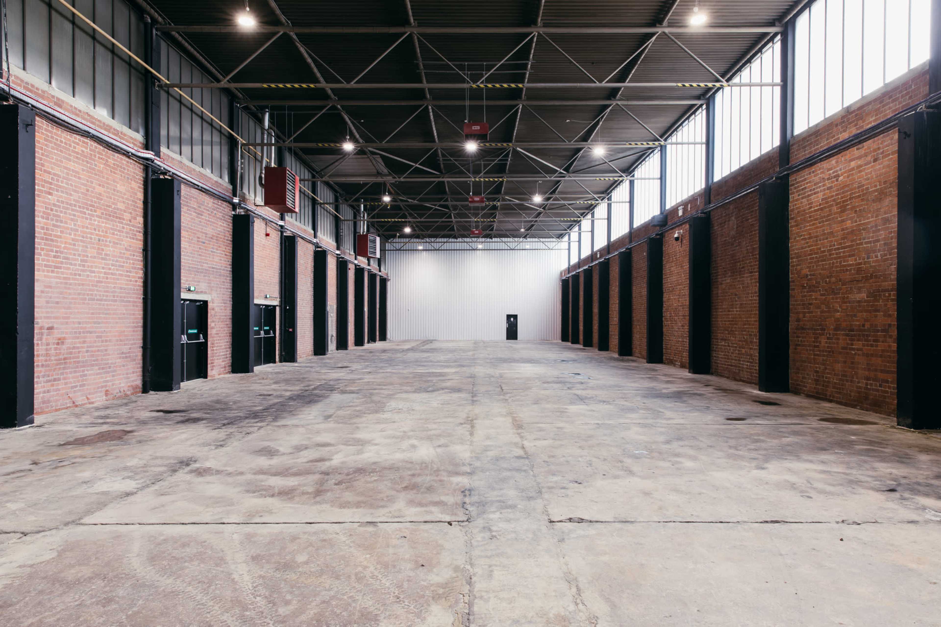 An empty industrial warehouse with exposed brick walls, large steel columns, and overhead lighting.