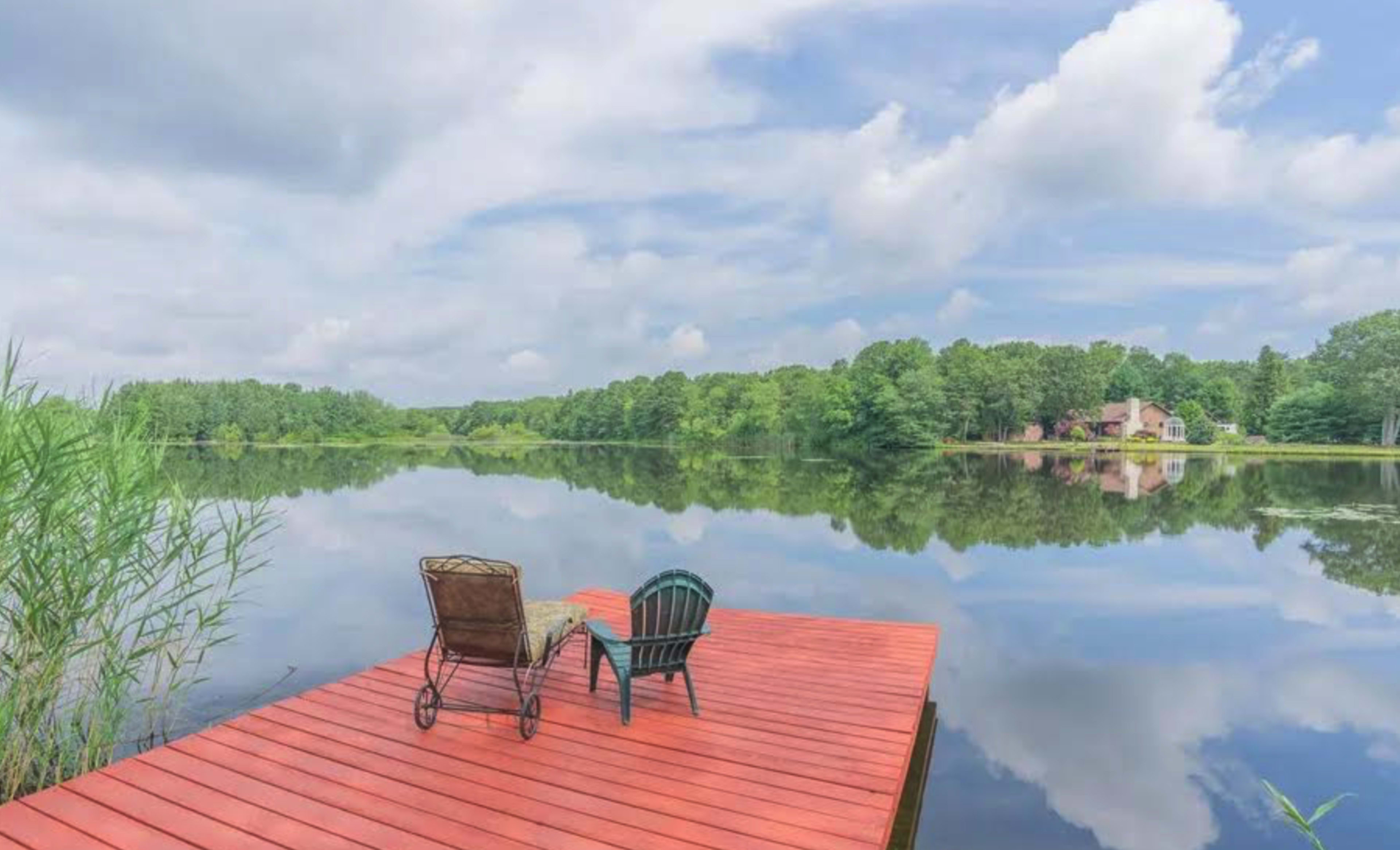 A red wooden dock extends over a calm lake, with a chair and a small table positioned on it, surrounded by lush greenery and reflections in the water.