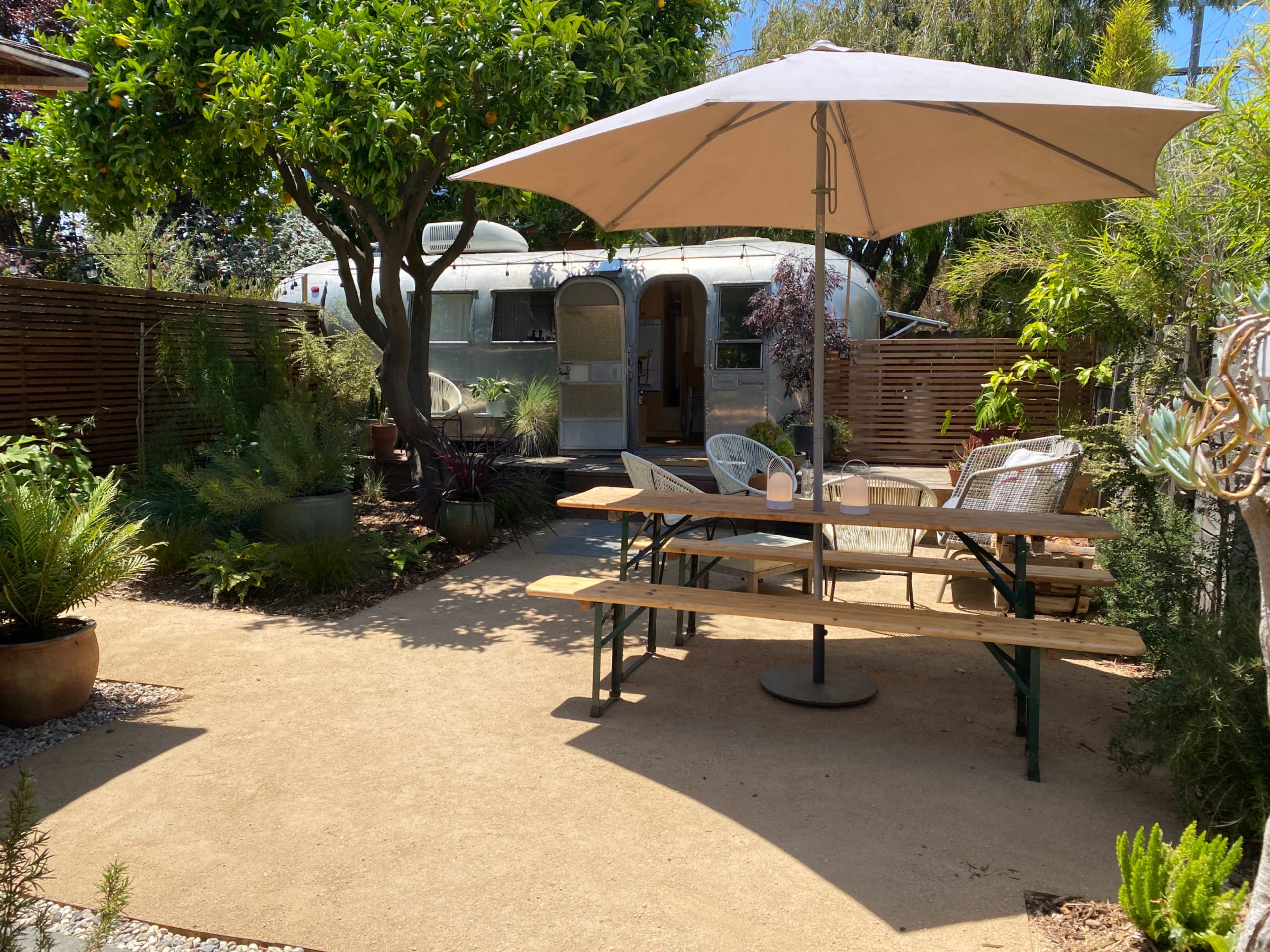 A backyard patio features a wooden table under a large umbrella, surrounded by potted plants and a vintage trailer in the background.