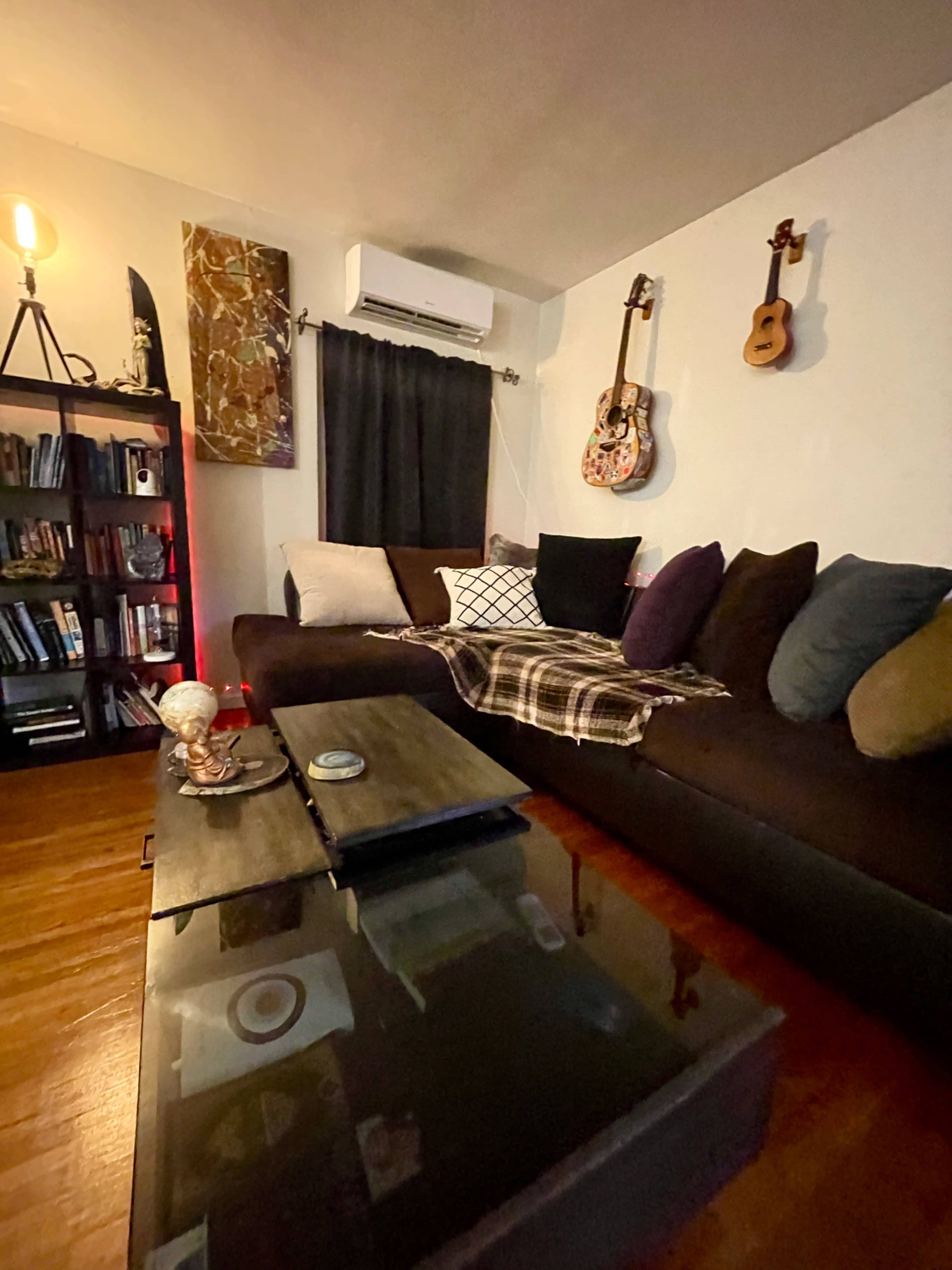 The image shows a cozy living room with a dark sectional sofa, a wooden coffee table, a bookshelf, and decorative items including a couple of guitars on the wall.