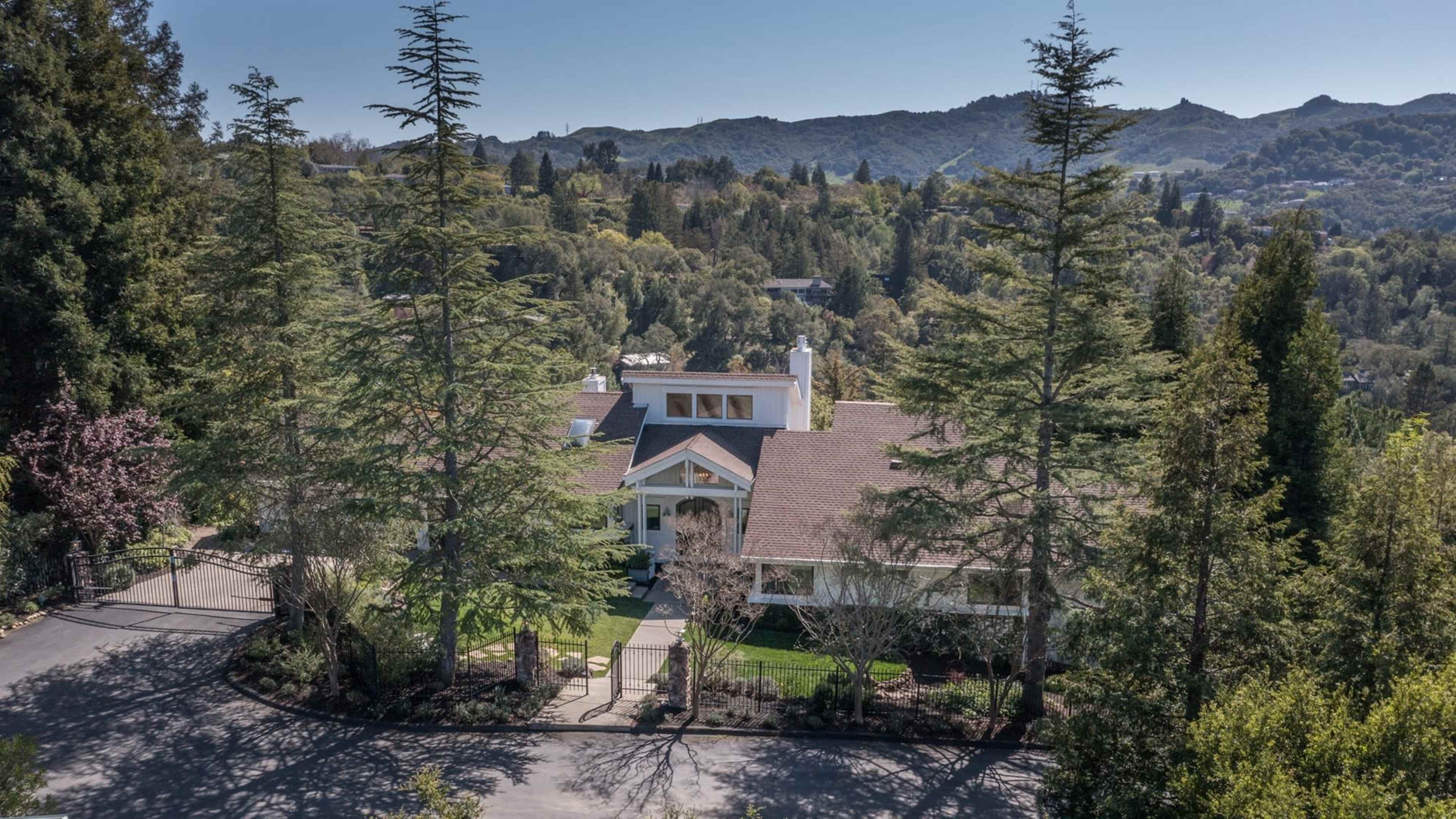 A spacious house is surrounded by tall trees and greenery, set against a backdrop of distant hills.