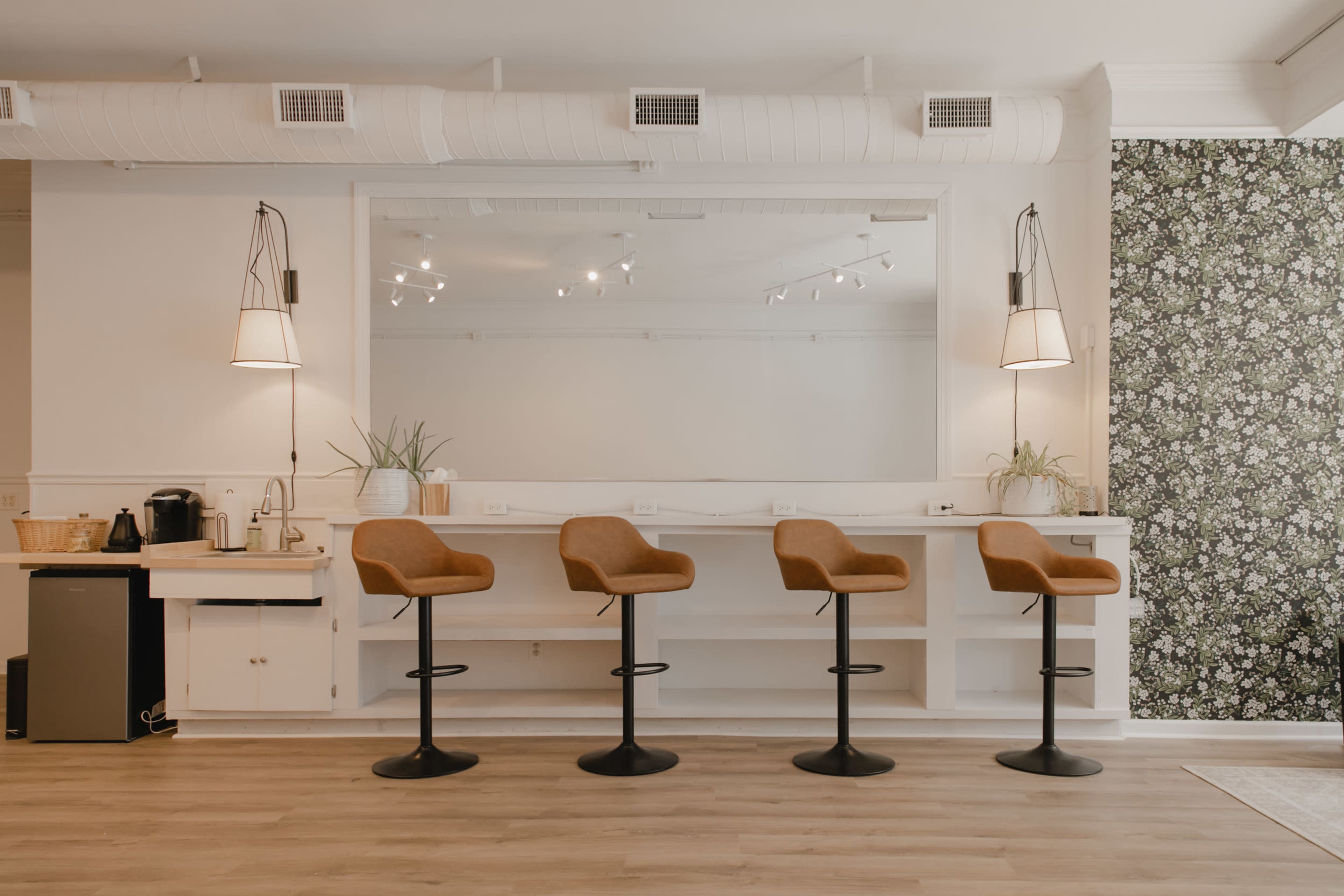 The image shows a modern interior space with a row of four brown bar stools in front of a white counter and a large mirror, flanked by wall-mounted light fixtures and a floral wallpaper.