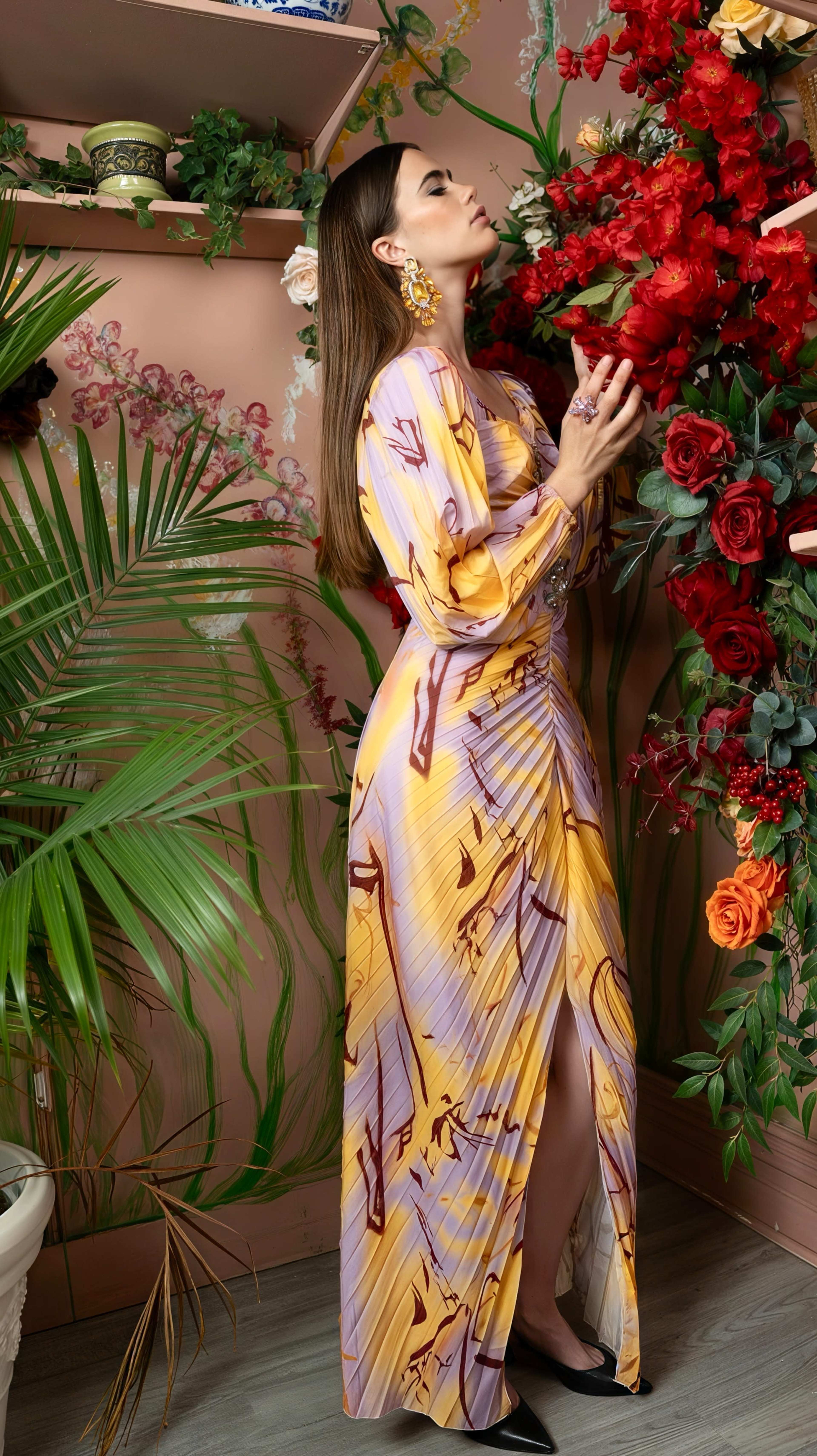 A woman in a colorful patterned dress stands beside a wall decorated with large red flowers and green plants.