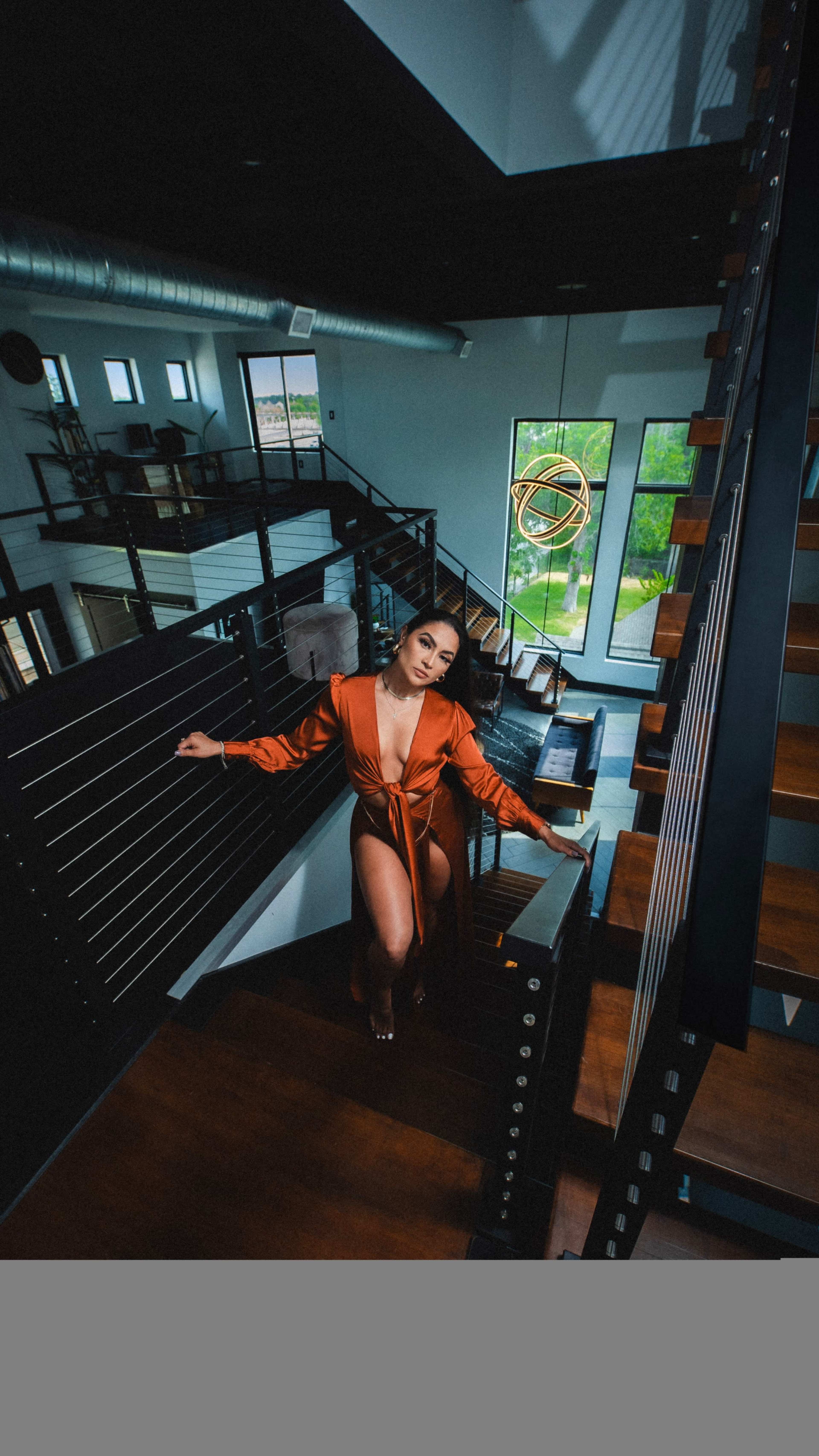 A woman in an orange dress poses on a staircase in a modern, well-lit building with large windows.
