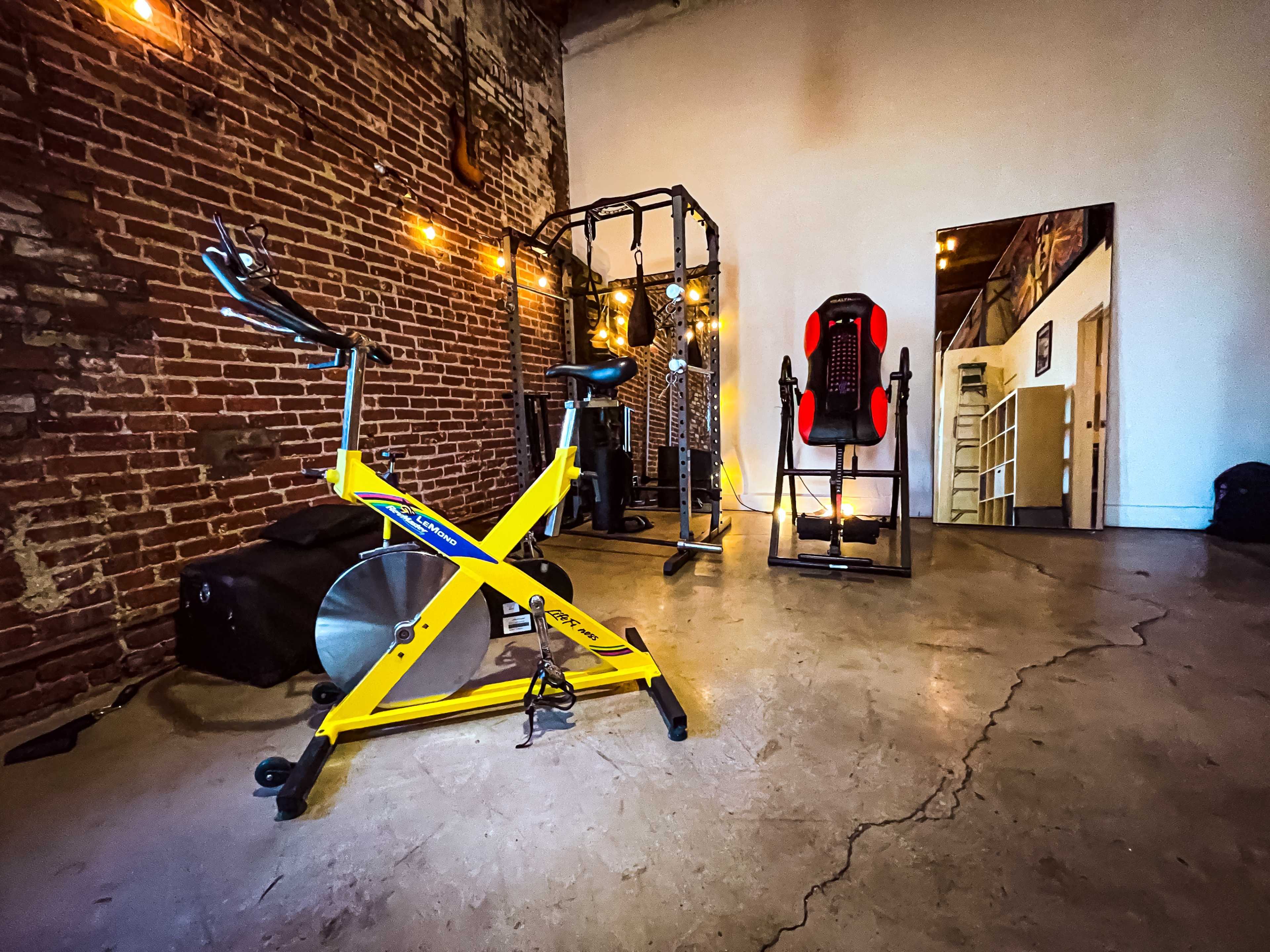 A fitness space featuring yellow exercise equipment, a multi-station gym setup, and a mirrored wall against a brick backdrop.