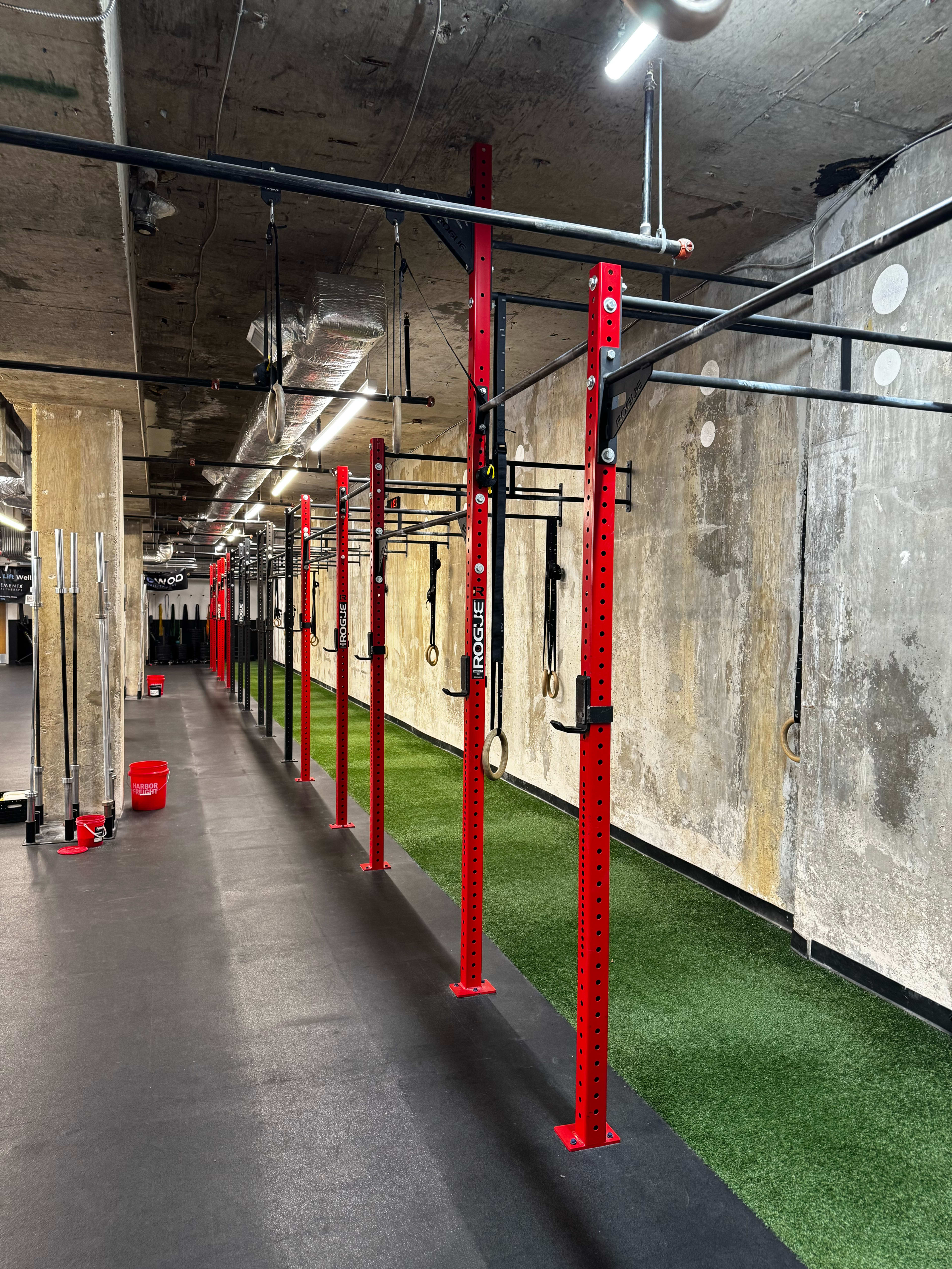 The image shows a gym interior with red squat racks and pull-up bars arranged along a black rubber floor, with a section of green turf on one side.