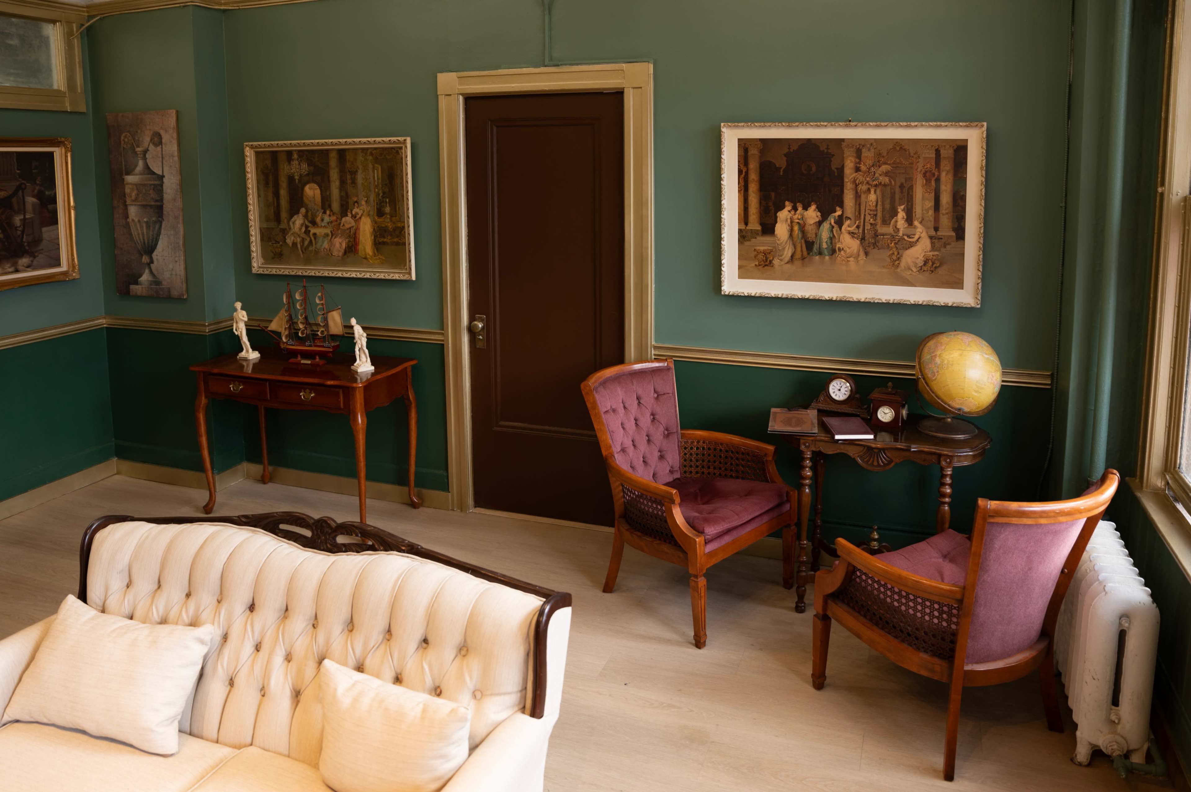 A vintage-style room with green walls, a white upholstered sofa, two wooden chairs, a small table with a globe, and framed artwork on the walls.