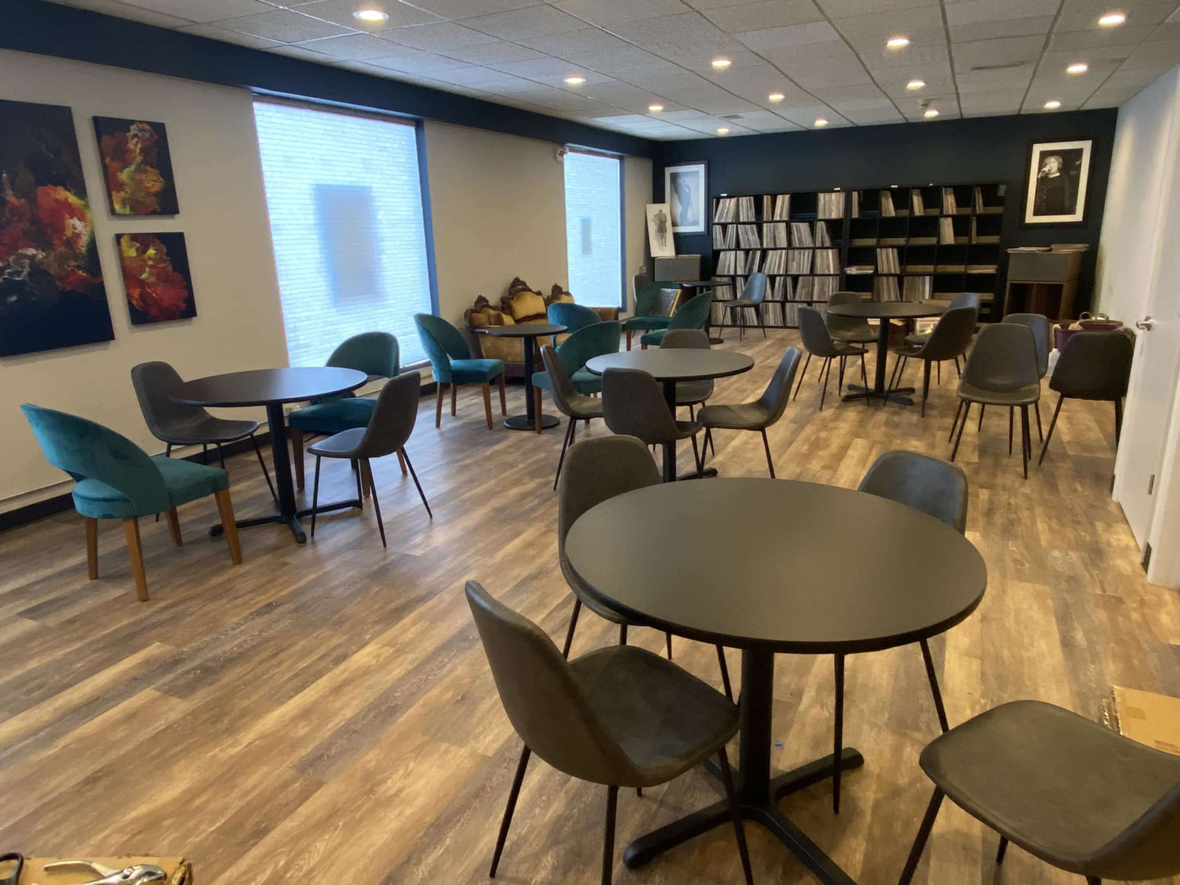 A modern room with several round tables and chairs, featuring artwork on the walls and a bookshelf filled with records.