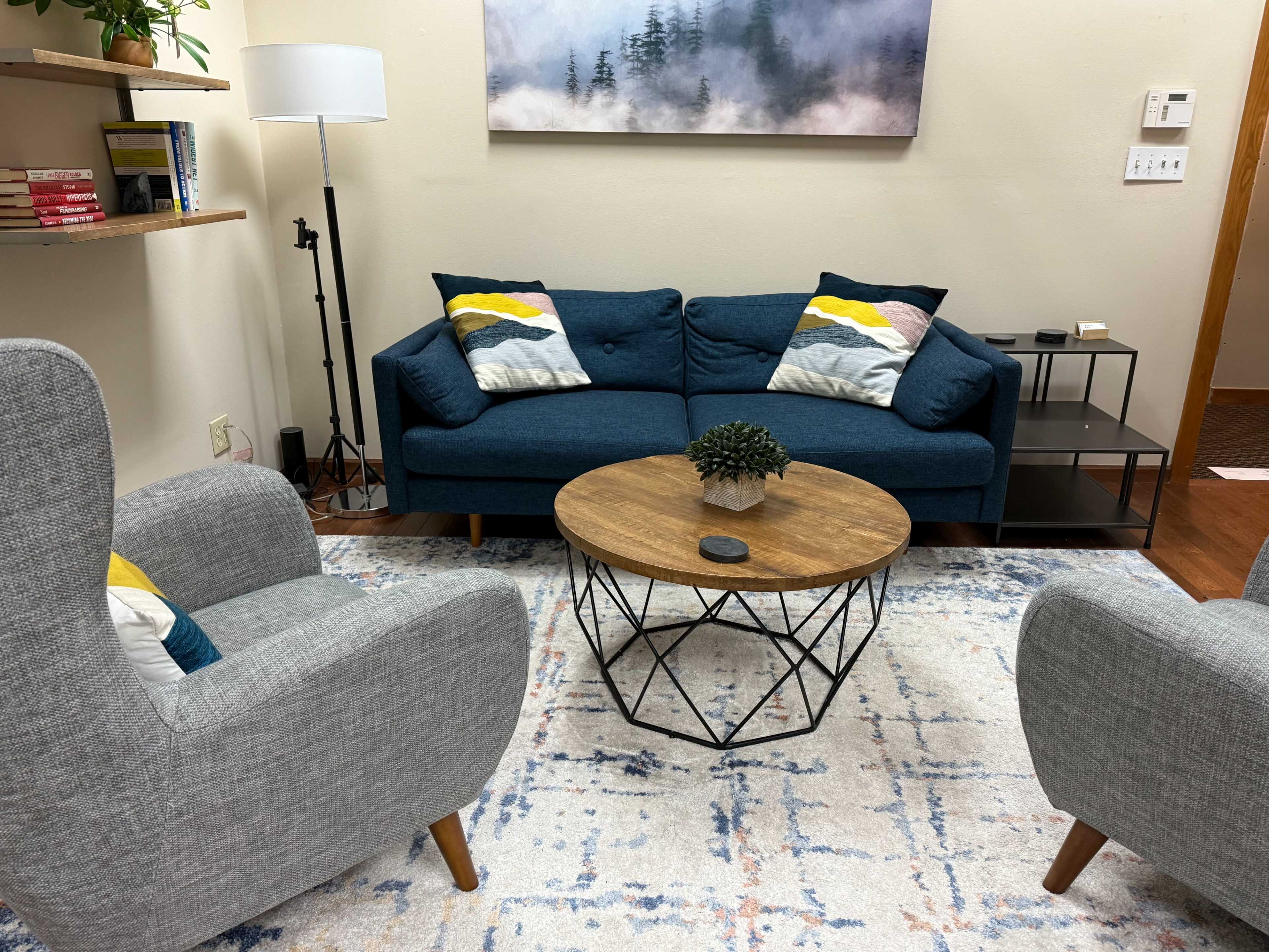The image shows a cozy living room setup featuring a blue couch with decorative pillows, two gray armchairs, a round wooden coffee table, and a shelf with books and plants.