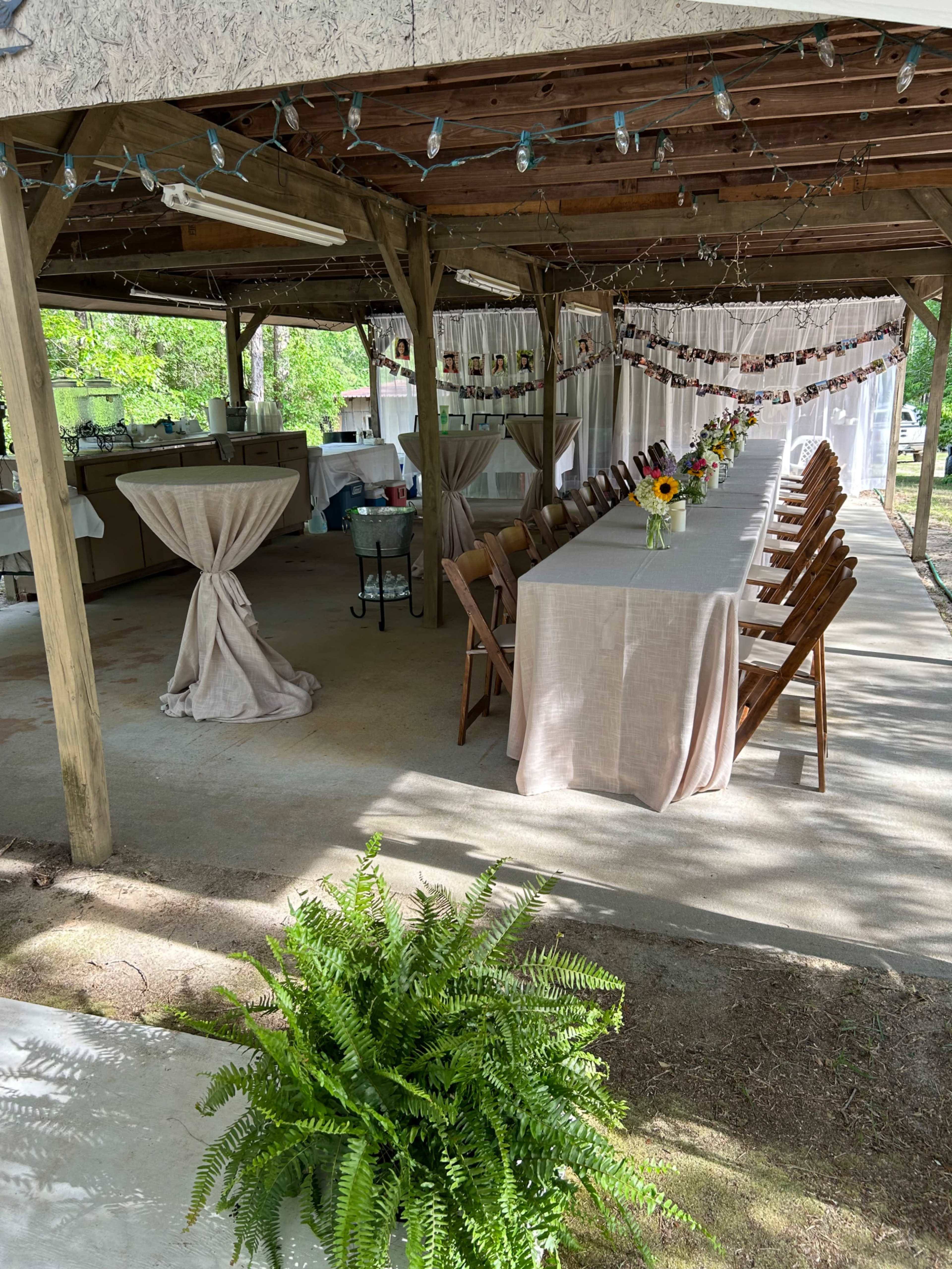 A covered outdoor space with a long table set for a gathering, surrounded by chairs and decorated with lights and flowers.
