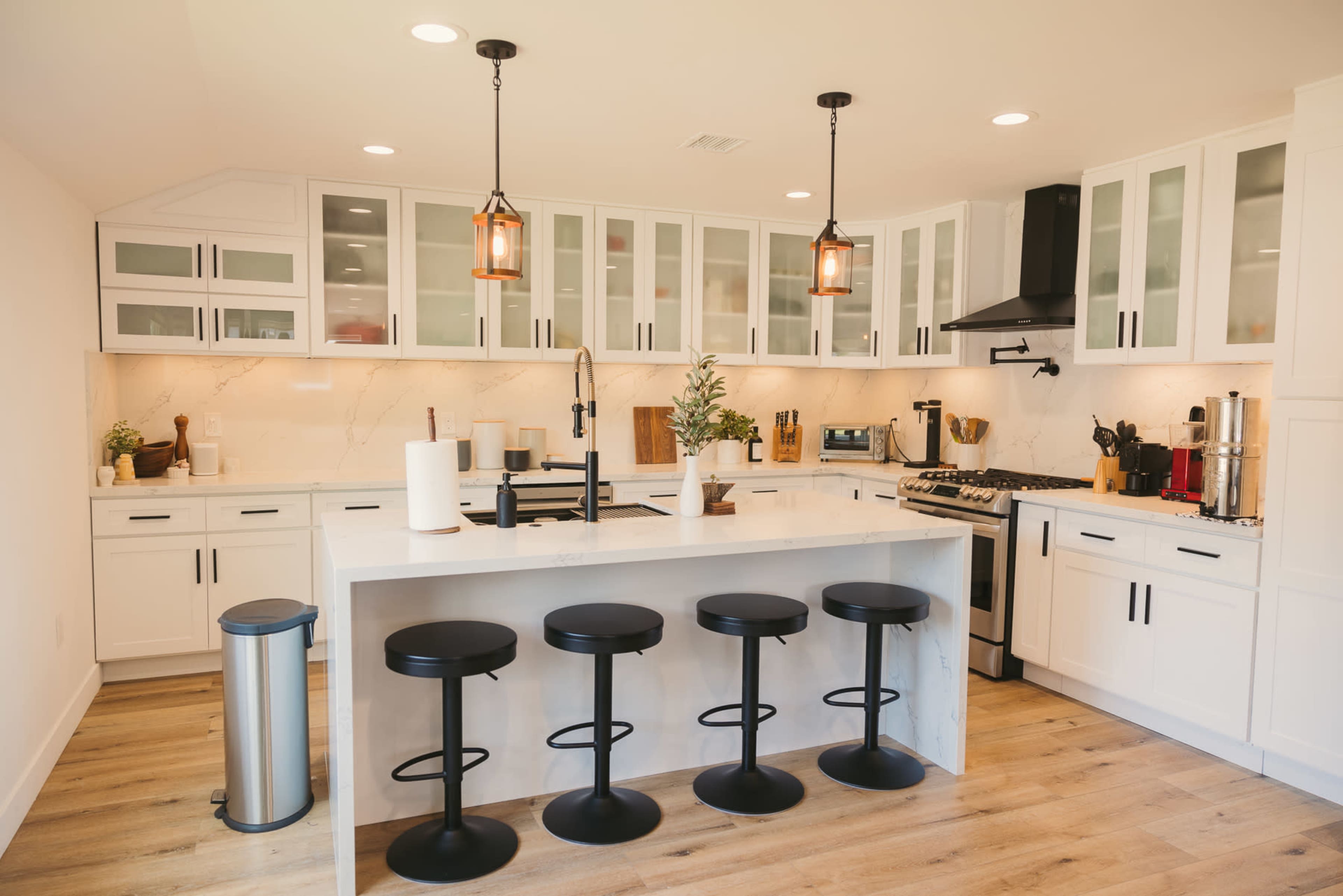 A modern kitchen features a central island with four black bar stools, white cabinetry, and stainless steel appliances.