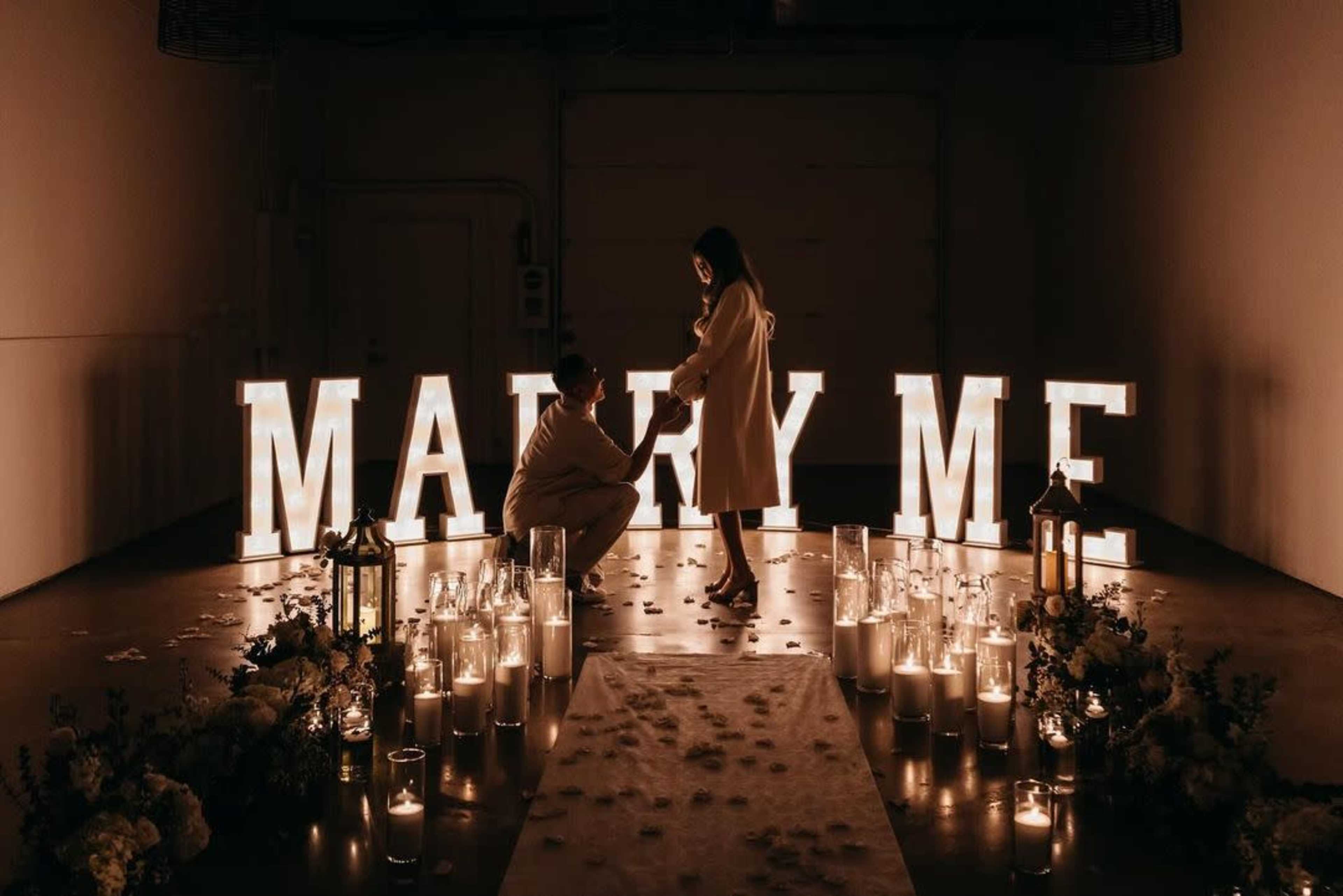 A person is kneeling in front of another near large illuminated "MARRY ME" letters, surrounded by candles and flowers in a dimly lit space.