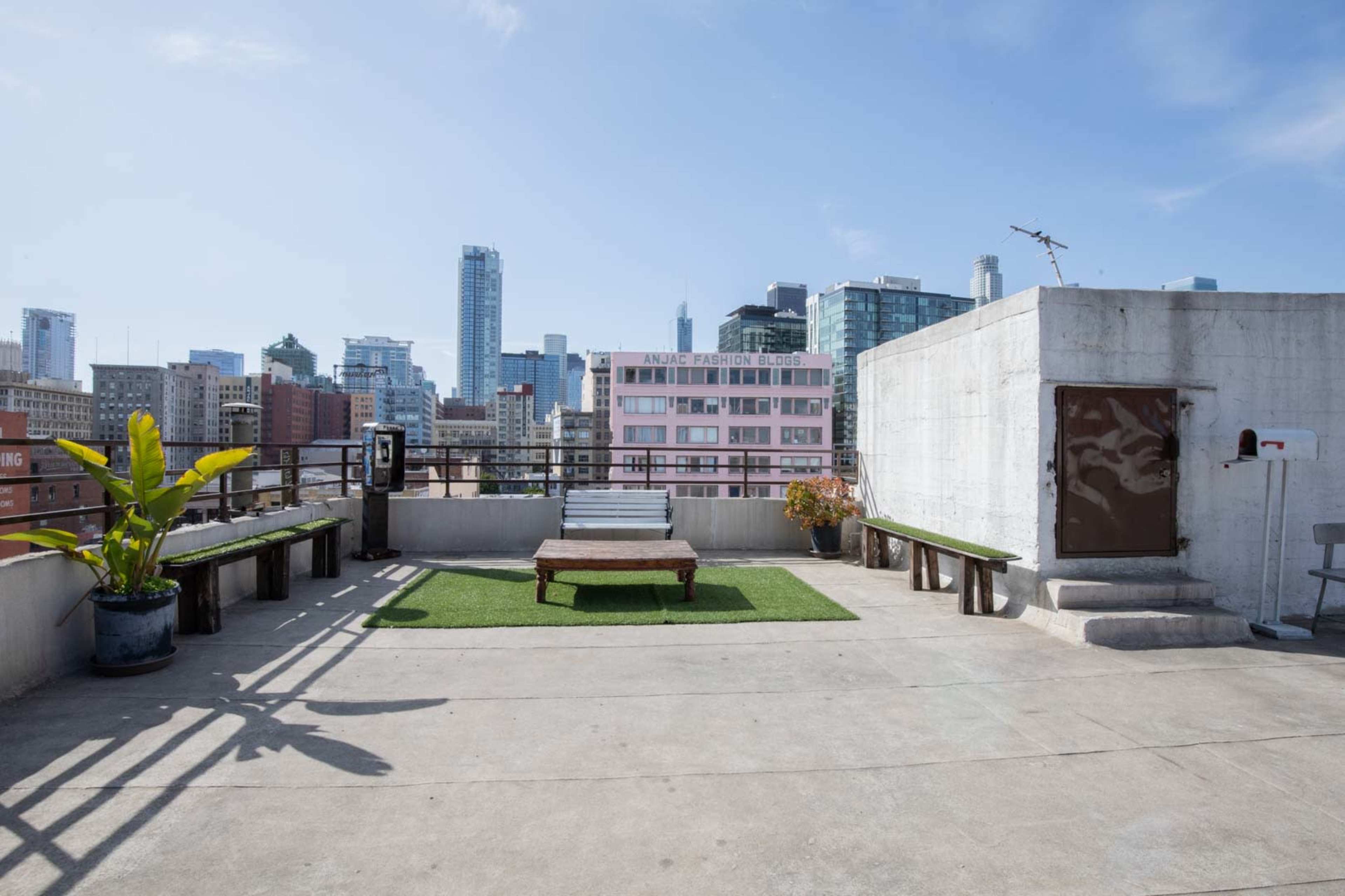 A rooftop space features a wooden platform surrounded by city skyline views and a few potted plants.