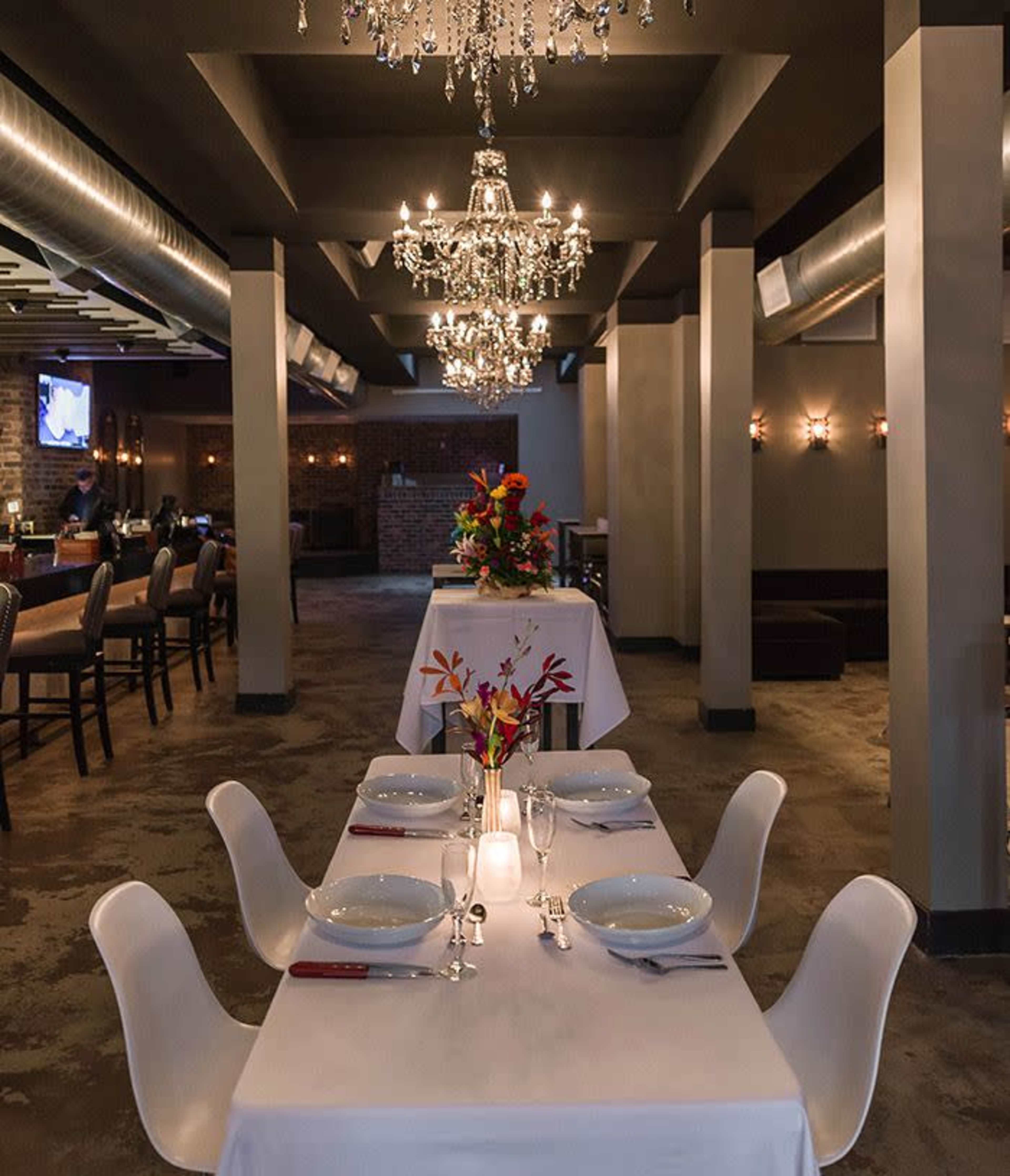A modern dining area features white tablecloths on tables adorned with floral centerpieces, surrounded by sleek chairs and illuminated by chandeliers.