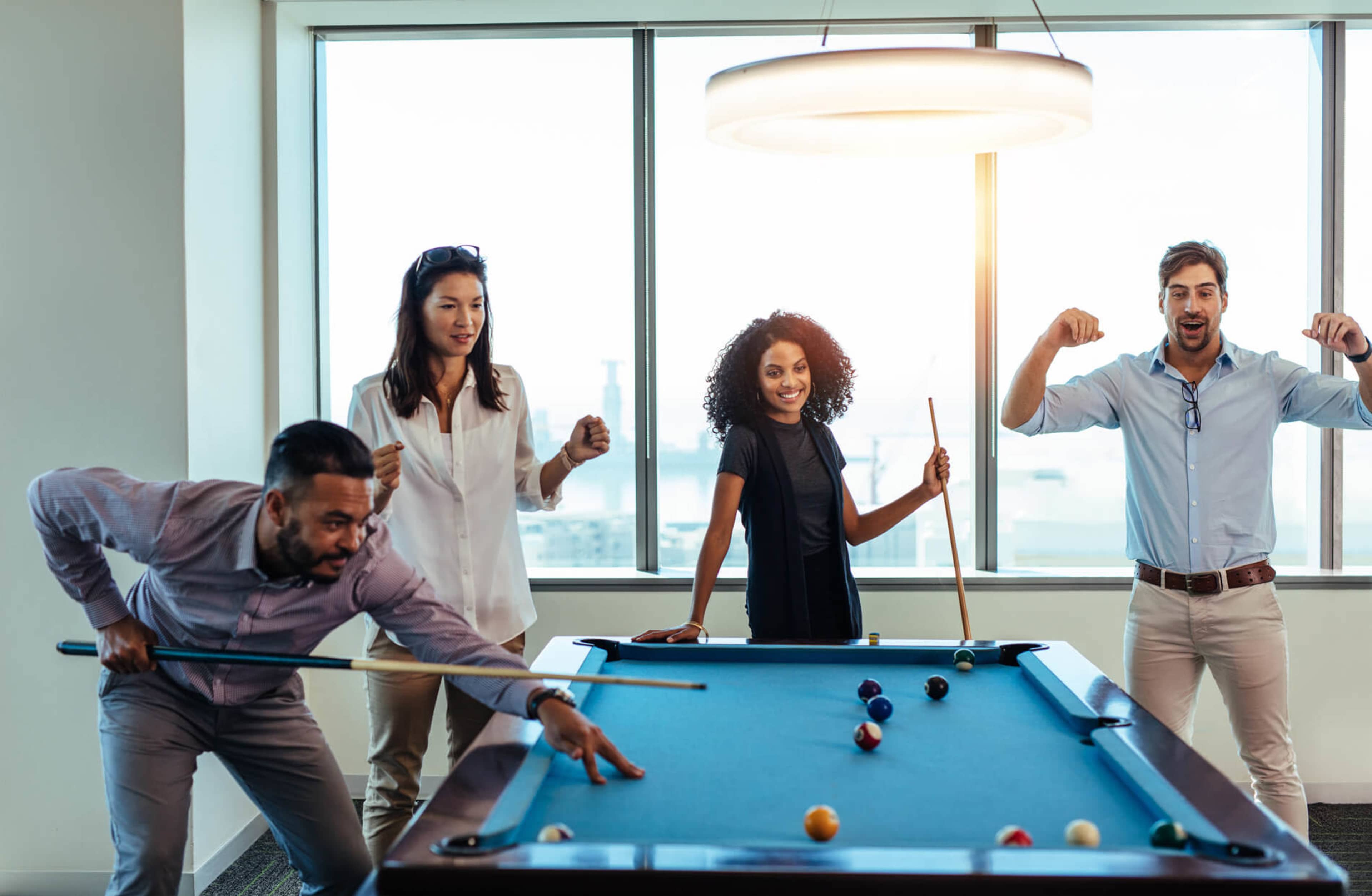 Four people are playing billiards in an office with large windows in the background.
