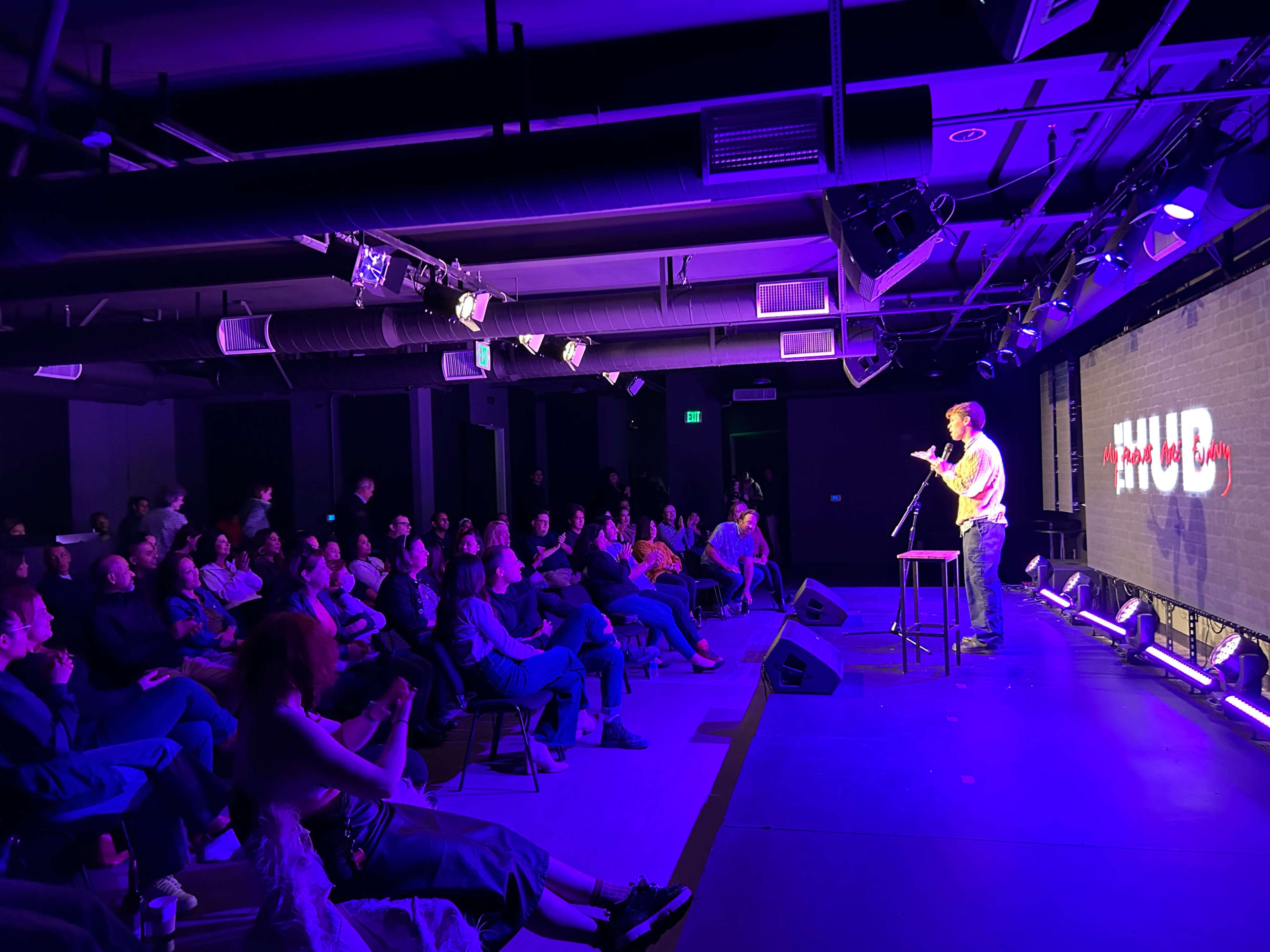 A comedian performs on stage in a dimly lit venue while an audience sits in chairs, facing the performer.