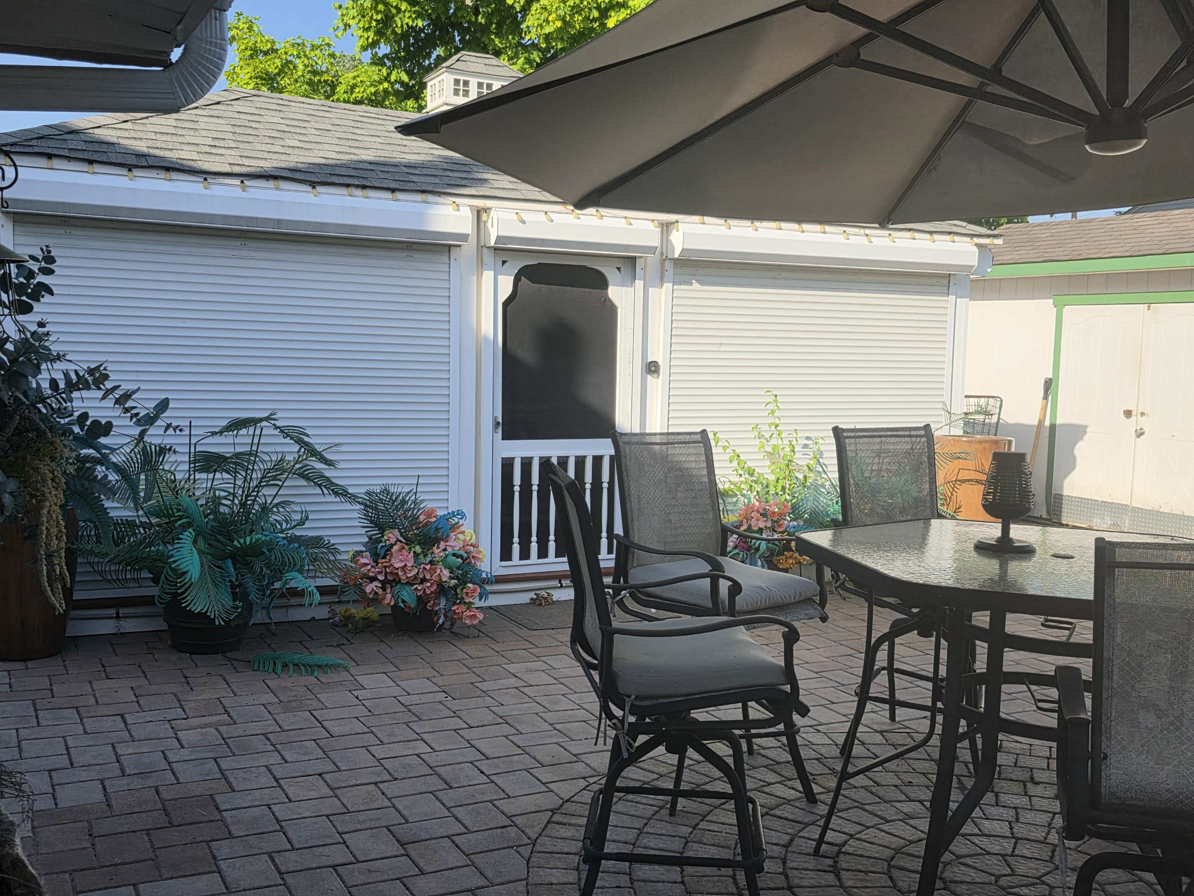 A patio area features a table and chairs, surrounded by decorative plants and with a shed visible in the background.